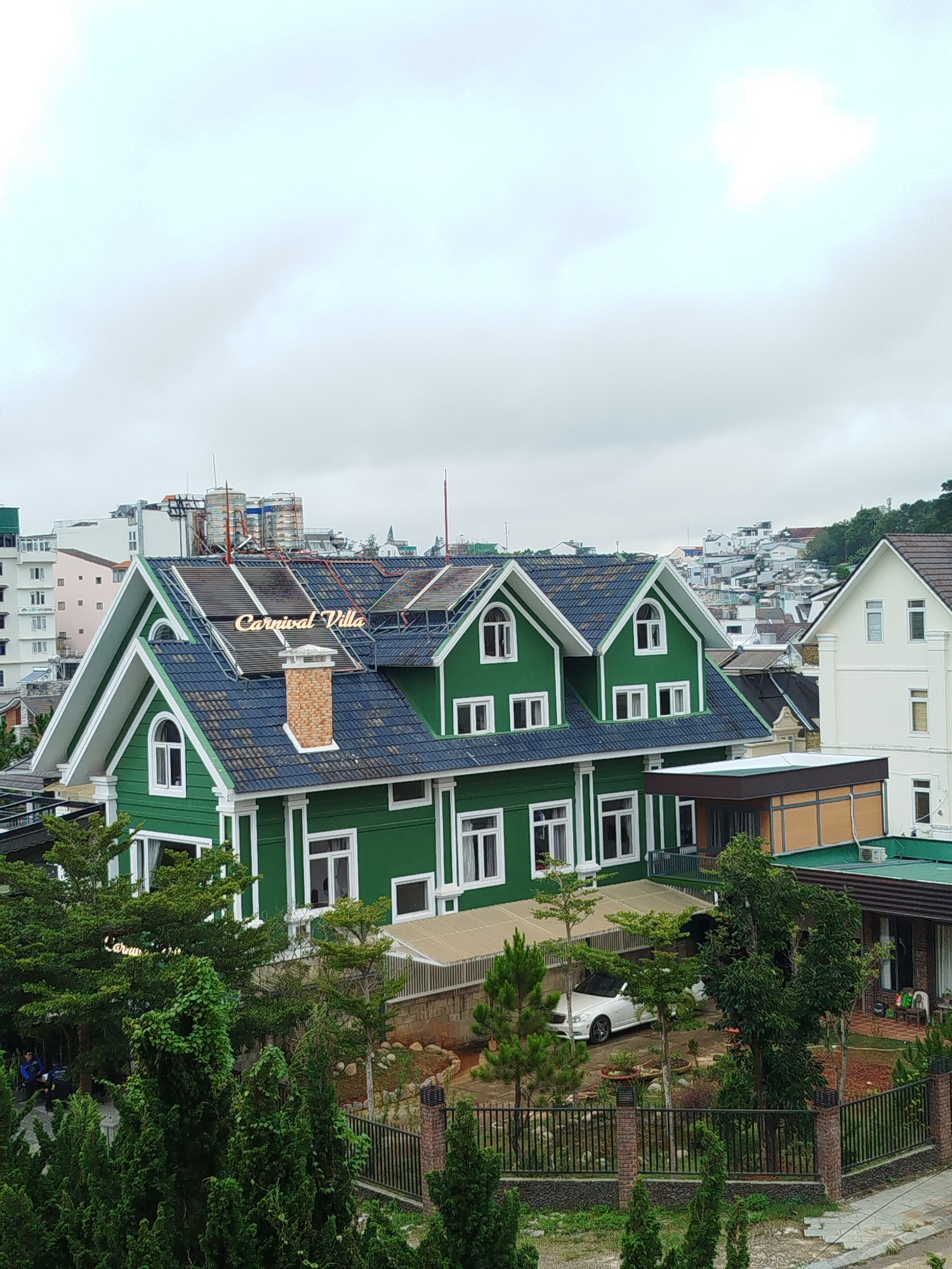 The image shows a green villa with a dark blue tiled roof, featuring white trim and multiple gabled windows. A brick chimney is visible on the roof, and a glowing sign that reads "Carnival Villa" is positioned on the roof's peak. The villa is surrounded by lush green trees and a black metal fence. In the background, there are other buildings, including a white house with a brown roof and several multi-story buildings. A white car is parked in the driveway of the villa. The sky is overcast. The scene is situated in Da Lat, Vietnam.