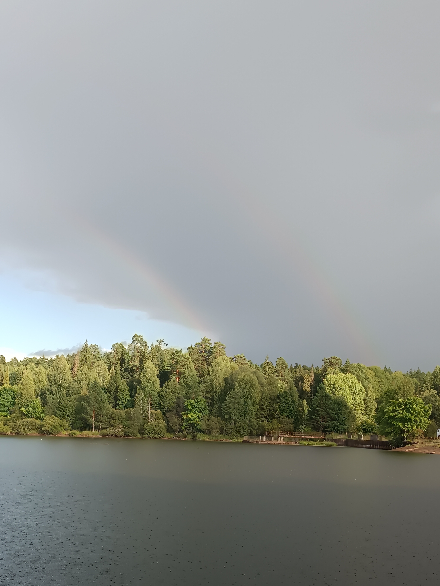 The image depicts a tranquil natural scene with a lake in the foreground and a dense forest lining the opposite shore. The sky is overcast, with two faint rainbows visible in the distance, suggesting recent or ongoing rainfall. The lake's surface is disturbed by raindrops, creating a speckled effect. Along the shoreline, a wooden pier extends into the water, and a small white structure, possibly a boathouse or shelter, is visible near the pier. The forest is lush and green, with a variety of trees. The overall impression is one of peaceful seclusion and natural beauty. While the image does not explicitly show any landmarks or identifying features of Chaikovsky, Russia, such a landscape is consistent with the natural surroundings of a city situated on the Kama River, known for its forested areas and water bodies.
