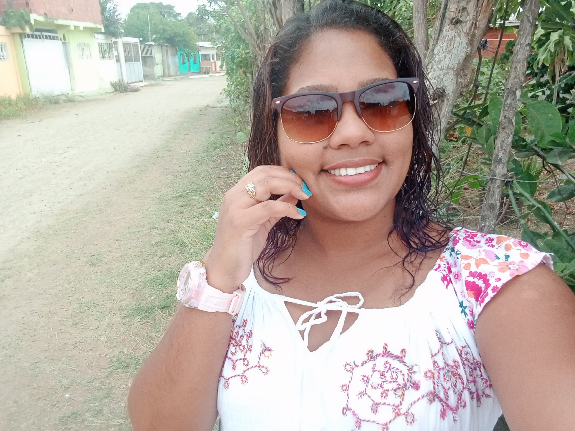 A dark-haired woman wearing sunglasses, a white floral top, and a pink watch stands in a grassy area next to a dirt road lined with single-story homes in Parroquia Ocumare, Venezuela. The woman is smiling and touching her face with her left hand, which has blue nail polish and a ring. Trees and bushes are visible behind her to the right.