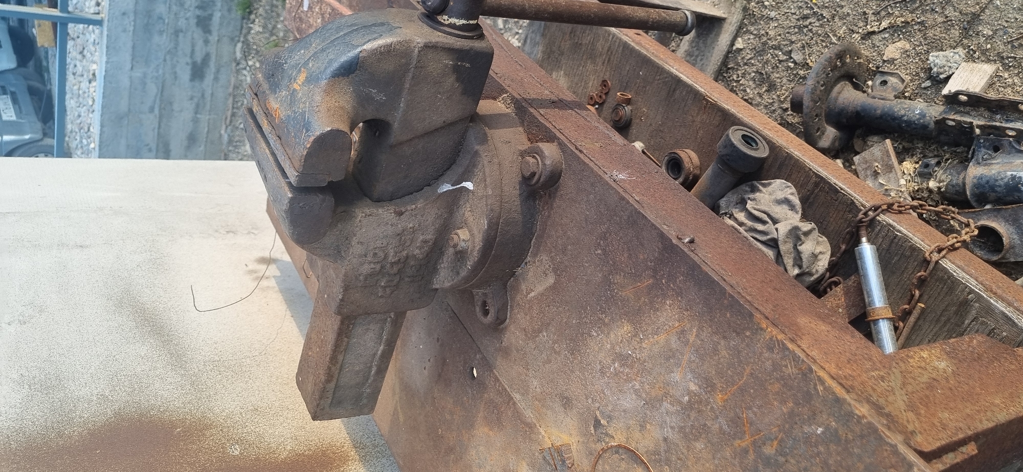 The image presents a close-up view of various old, heavily rusted metal objects and mechanical parts, likely in an outdoor industrial or storage setting. The primary subject is a substantial, rectangular metal frame or chassis, severely corroded and reddish-brown from rust.
A large, heavy-duty cast-iron vise, also showing significant rust and some chipped black paint on its moving jaw, is bolted onto the side of this metal frame. The vise's jaws are open, and its handle extends upwards and to the right. Below the vise, a light-colored concrete surface is visible, with a loose piece of dark wire on it.
Inside the hollow space of the main metal frame, an assortment of smaller items can be seen. These include several rusty metal nuts, washers, and bushings, as well as a crumpled piece of grey cloth. Further inside, a rusty metal chain is draped over a silver-colored, possibly hydraulic or pneumatic, cylinder.
In the background, beyond the metal frame, more old, black-painted and rusty metal components, possibly vehicle suspension parts or axles, lie on dirt and gravel. To the far left background, a blurry concrete wall or structure and part of a light-colored vehicle are faintly visible.
The scene is brightly lit by natural daylight, casting distinct shadows and suggesting sunny weather. The overall impression is one of disuse and weathering. No legible text or markings are visible on any of the objects. The location context indicates this scene is in Ленинское сельское поселение, Ukraine.