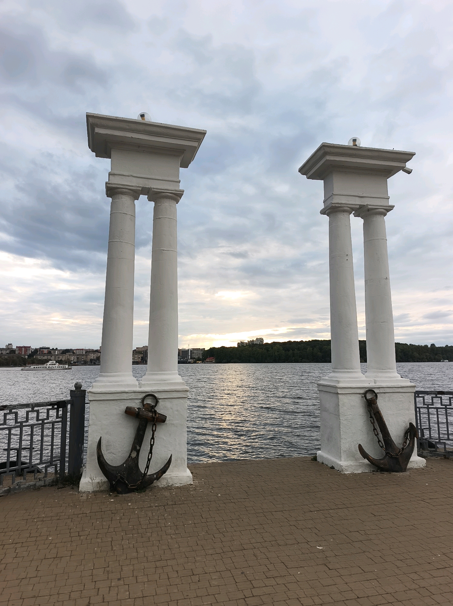 Two white stone columns, topped with square pedestals and decorative lights, stand on a brick-paved embankment overlooking a body of water. Each column has an anchor and chain attached to its base. The water is calm with ripples reflecting the overcast sky. In the distance, a treelined shore with buildings and a boat are visible. The sky is cloudy with patches of light breaking through.