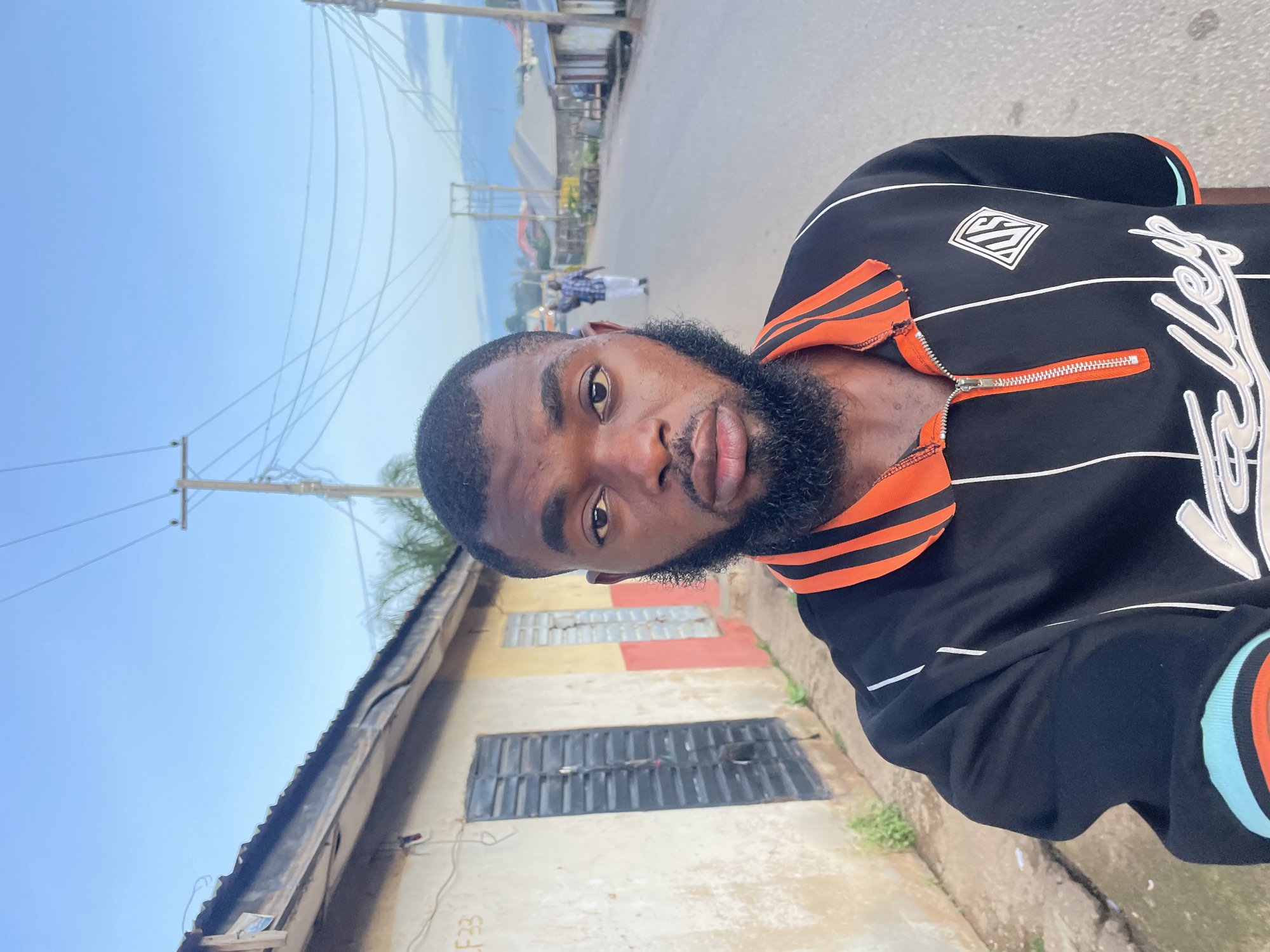 A man with a full beard takes a selfie on a street in Jos, Nigeria. He is wearing a black and orange collared shirt with white stripes and lettering. Behind him, a street stretches into the distance, lined with buildings and utility poles. The sky is a pale blue with some clouds. The man is the primary subject, with the urban environment serving as the background. The overall scene depicts a casual, everyday moment in an African city.