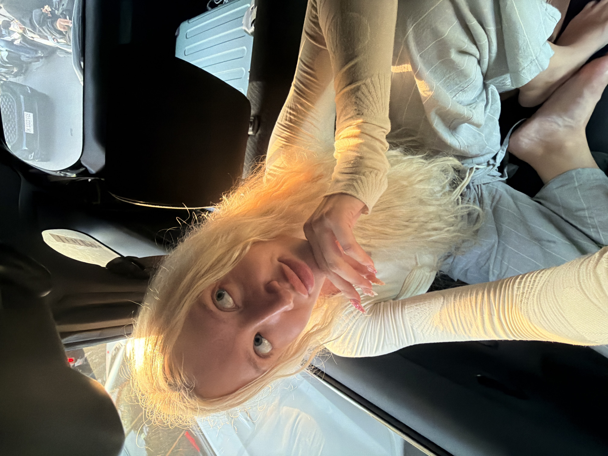 A young Caucasian woman with long blonde hair is sitting in the back seat of a car, possibly in North Kuta, Indonesia. The sun is shining brightly, casting a warm glow on her face and hair. She is wearing a white long-sleeved shirt and grey striped pants. Her left hand is resting on her chin, and she is looking off to the side. The interior of the car is visible, including the headrest and the back of the seat in front of her. Outside the car, other vehicles and people can be seen, suggesting a busy urban environment. The license plate of a black SUV in front of the car is visible and reads "DK 1322 FCE".