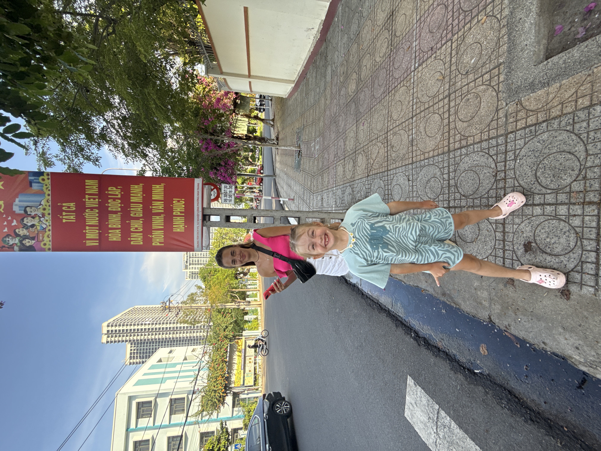 Two females, an adult and a child, stand on a sidewalk in Nha Trang, Vietnam. The adult, positioned behind the child, wears a bright pink off-the-shoulder top and a white skirt. She holds a pink smartphone in her right hand and a black crossbody bag is slung over her left shoulder. The child, in front and facing the camera, wears a light blue t-shirt with a zebra-like pattern and matching shorts. She has a white necklace with flower charms and is wearing pink Crocs.
Behind them, a large red banner with Vietnamese text and images is displayed vertically. The banner features pictures of people and sunflowers. The surrounding environment includes a city street with a black car and a motorcycle visible, tall buildings in the background, trees, and a sidewalk made of patterned tiles. A street light and a bus stop sign are also present. The scene is captured during daylight.