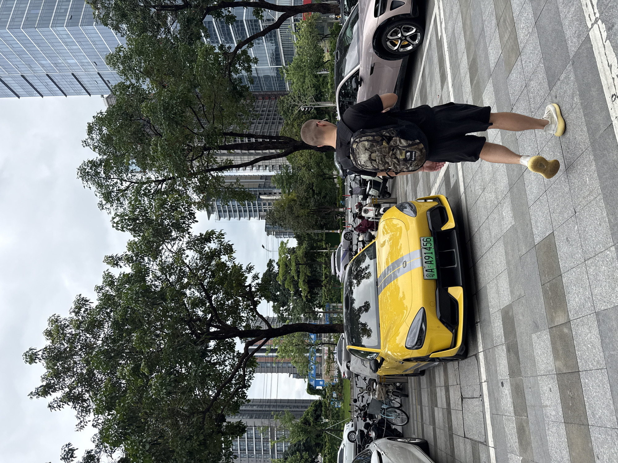 In Tianhe District, China, a young man with short hair and a camouflage backpack walks away from the camera on a gray tiled sidewalk. He wears a black t-shirt and black shorts, and his right leg is slightly bent as he walks. In front of him, a bright yellow sports car with gray racing stripes is parked on the right side of a paved area. The car has a license plate that reads "A-A91456". To the right of the yellow car is a light pink sedan. Beyond the cars, a line of trees with full green foliage runs parallel to the street. In the background, several modern high-rise buildings dominate the skyline. The sky is overcast. Several motorcycles are parked behind the yellow car.
