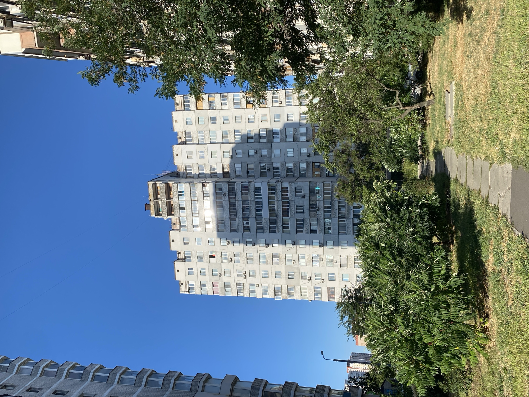 A tall, concrete apartment building dominates the center of the frame, its facade marked by numerous windows and air conditioning units. A shadow from an unseen object falls across the building. To the left, another apartment building, constructed with grey bricks, is partially visible. In the foreground, a stone pathway winds through a patch of dry, yellowing grass and several leafy green bushes and small trees. The sky above is a clear, deep blue. In the distance, other buildings and a street lamp can be seen. The scene appears to be a residential area in a city, bathed in bright sunlight.