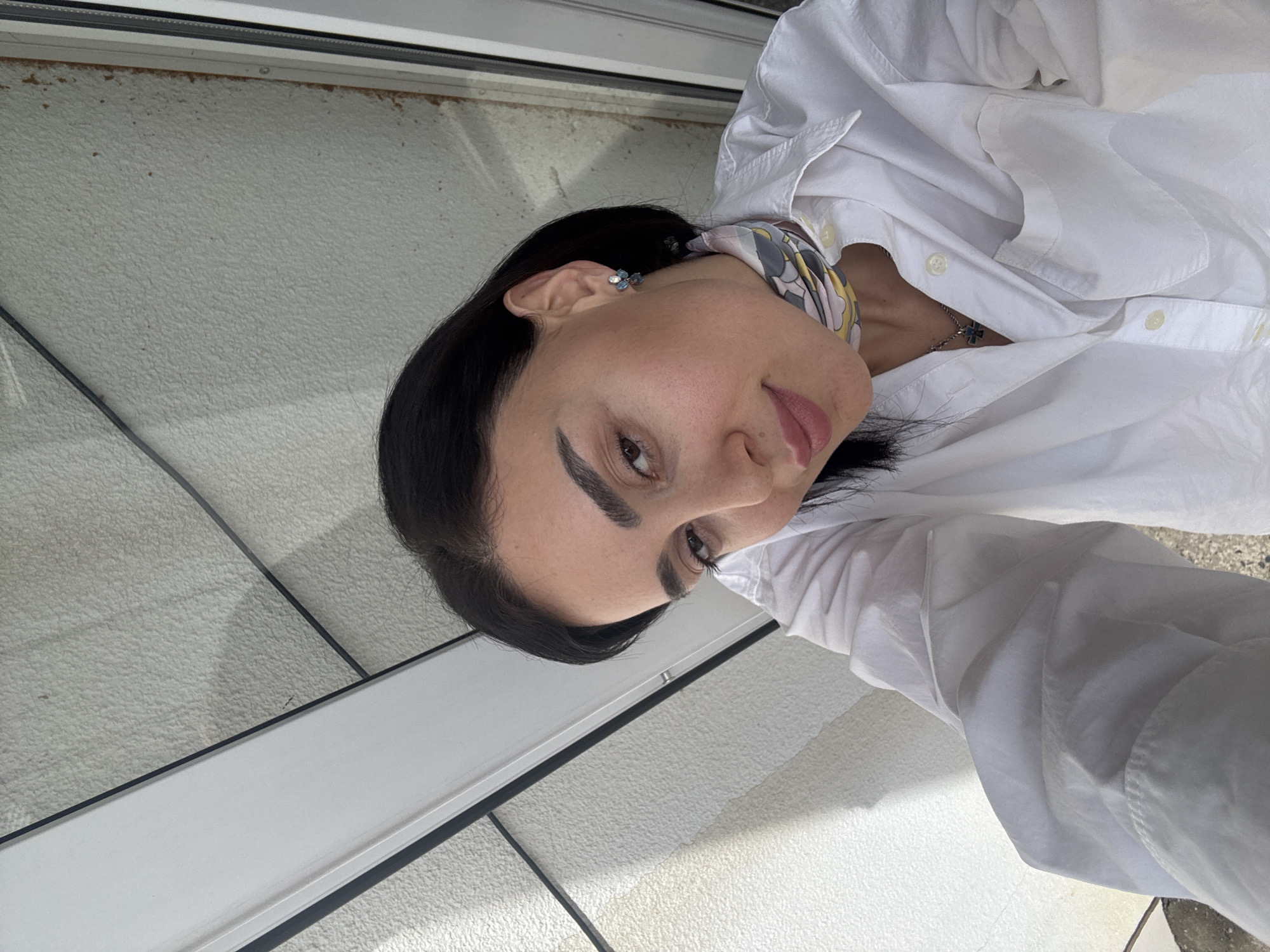 A woman with dark hair and prominent eyebrows is wearing a white shirt and a scarf with a yellow and gray pattern. She also has stud earrings and a silver necklace with a small cross. The location appears to be indoors near a window. The city is not discernible from the image.