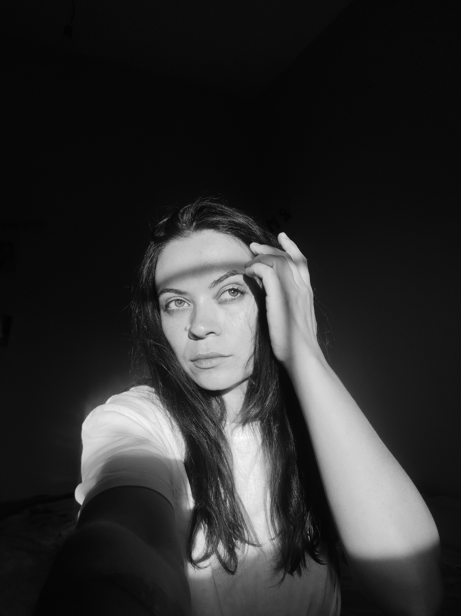 A black and white, close-up, self-portrait photograph captures a young adult female looking upwards and slightly to the left. The female has dark, long hair, a neutral facial expression, and is wearing a white t-shirt. Her right hand is raised near her forehead, with fingers spread, appearing to shield her eyes or pose. A strong, directional light source, likely sunlight, creates a prominent rectangular band of light across her forehead, left eye, and the top of her right hand, casting dramatic shadows. The light also illuminates her right arm and parts of her torso. The background is almost entirely black, indicating deep shadow and isolating the subject. The setting is indoors, in the city of Unknown, Unknown.