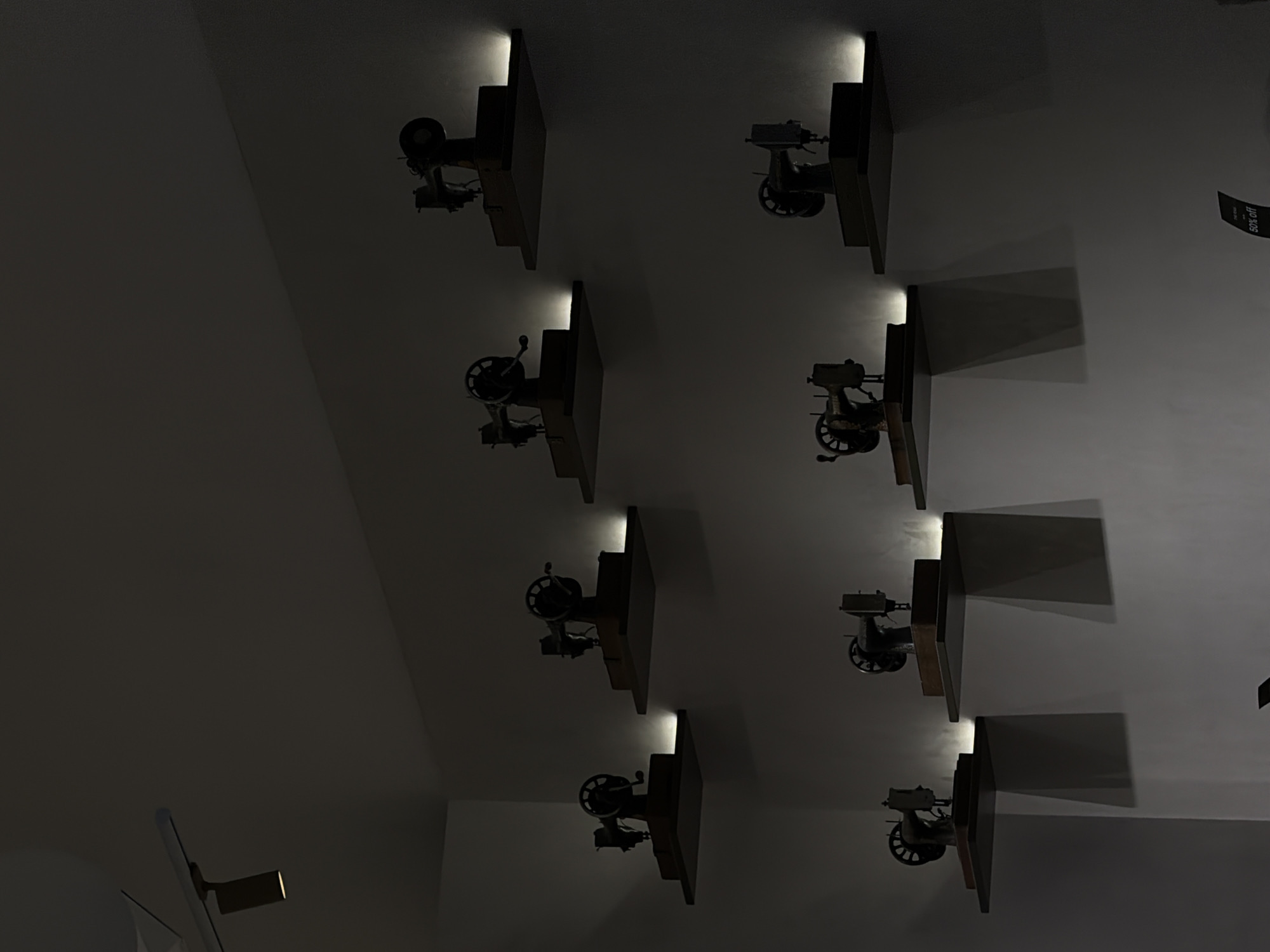 A group of antique sewing machines are displayed on shelves on a wall. There are eight sewing machines visible in the image, arranged in two rows of four. Each sewing machine is a miniature replica and is illuminated by a subtle light source from behind the shelf. The scene is dimly lit, creating a dramatic effect with shadows cast by the sewing machines and shelves. A spotlight is visible on the ceiling in the upper left corner of the image. The location is identified as Ramat Gan, Israel.