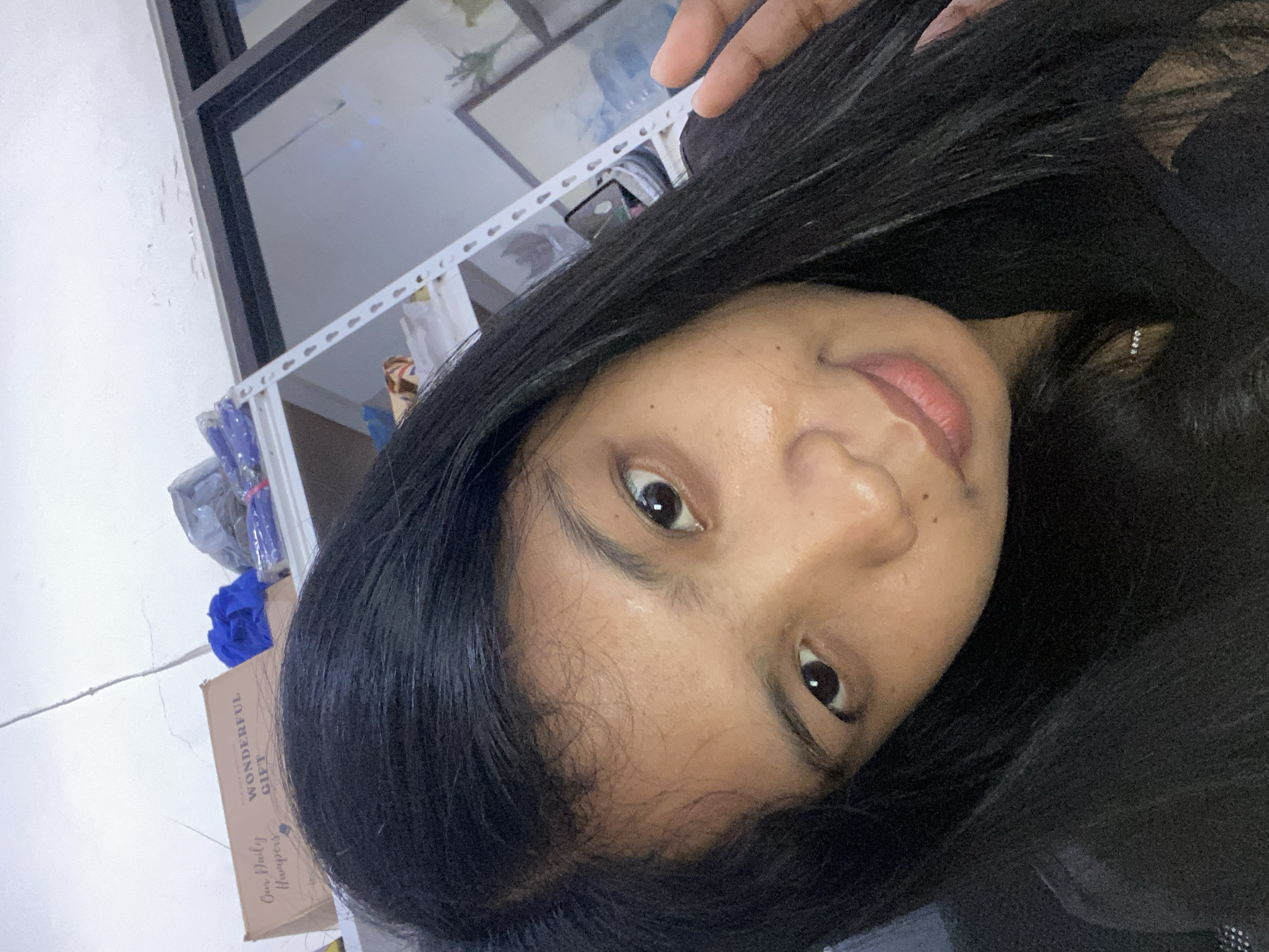 A selfie taken by a woman with long, black hair. The woman's face is the central focus, showing her eyes, nose, and lips. She is wearing an earring. In the background are a window, shelves with various items, a blue fabric, and a cardboard box labeled "Wonderful Gift". The location is Negara, Indonesia.