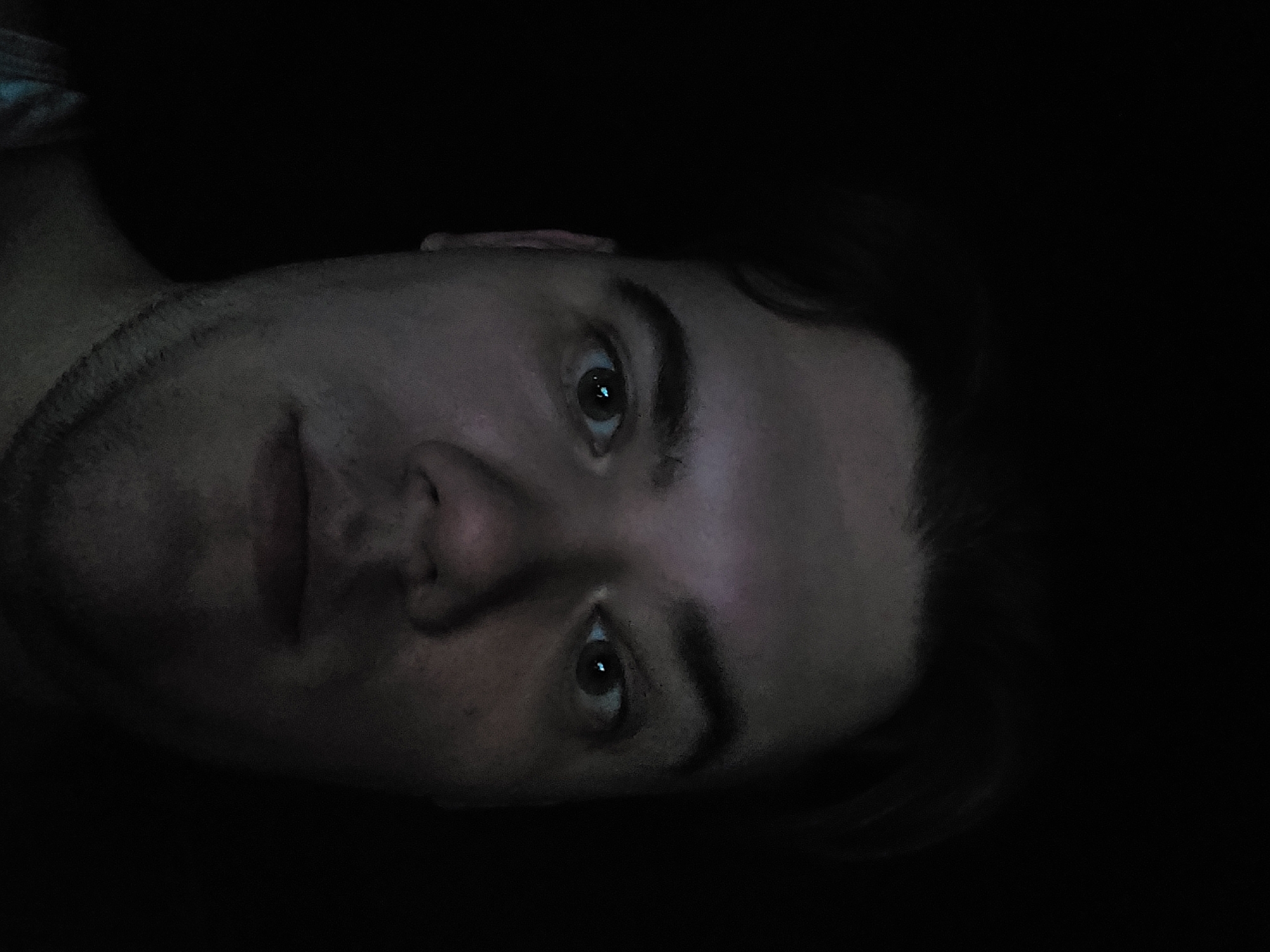 A dimly lit, close-up portrait of a man's face is displayed. The man has short dark hair, dark eyebrows, and brown eyes. He has stubble on his face. The background is mostly black, obscuring any details. The man's face is the primary subject of the image. There is no discernible location in the city of Tashkent, Uzbekistan, or any identifiable environment. The image predominantly focuses on the man's facial features.