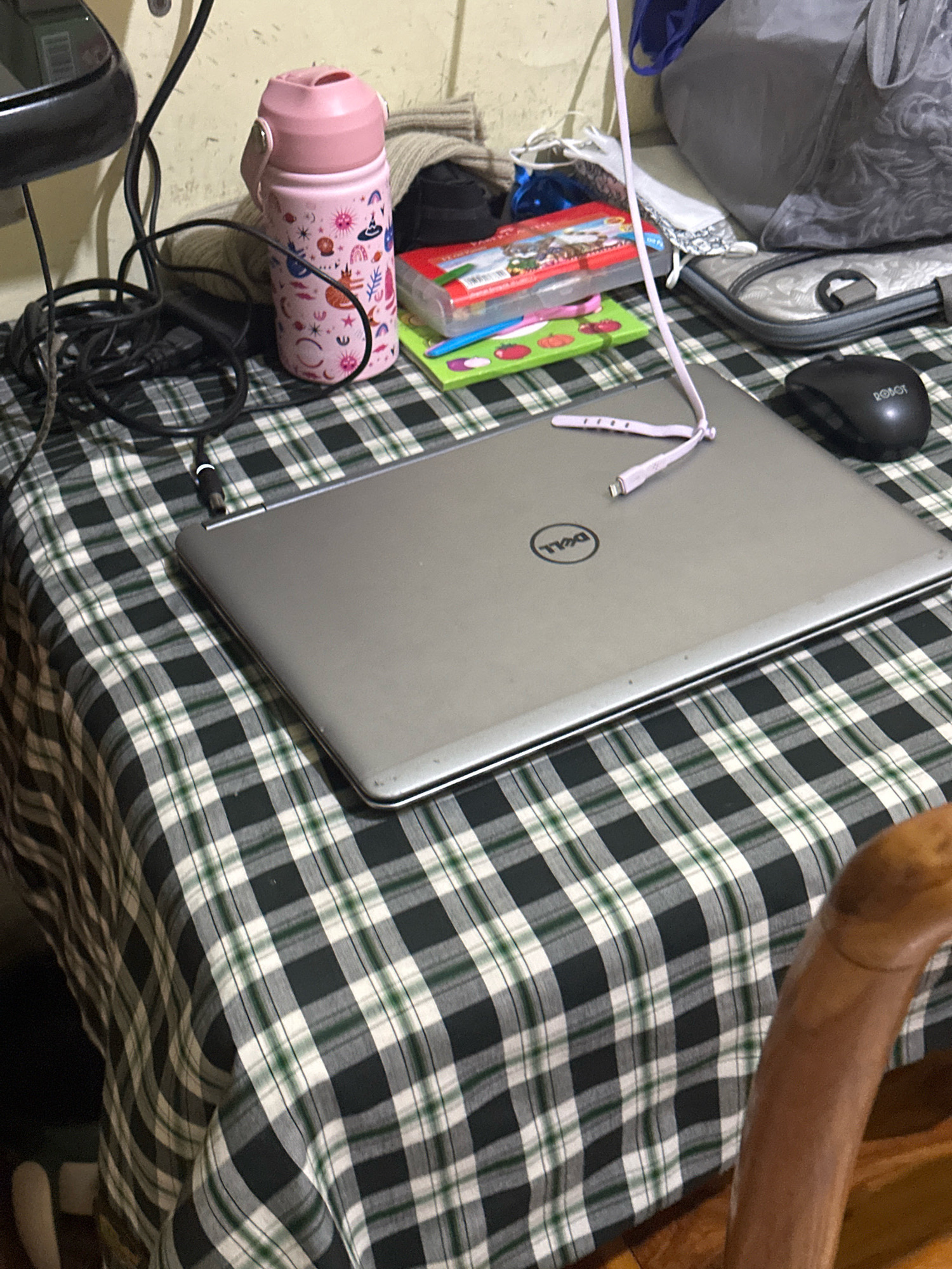 The image depicts a close-up, elevated view of a table covered with a green, white, and black plaid tablecloth. On the table, a closed silver Dell laptop is centrally positioned. To the left of the laptop, a black coiled power cord is visible, leading to an unspecified device. Next to the power cord and slightly behind it, a pink water bottle with a white cap and a decorative pattern is present. A light purple charging cable is draped over the laptop. To the right of the laptop, a black computer mouse is situated. In the background, the wall is a neutral color, and other objects such as a bag and a bundle of fabric are partially visible. The scene is domestic and appears to be indoors.