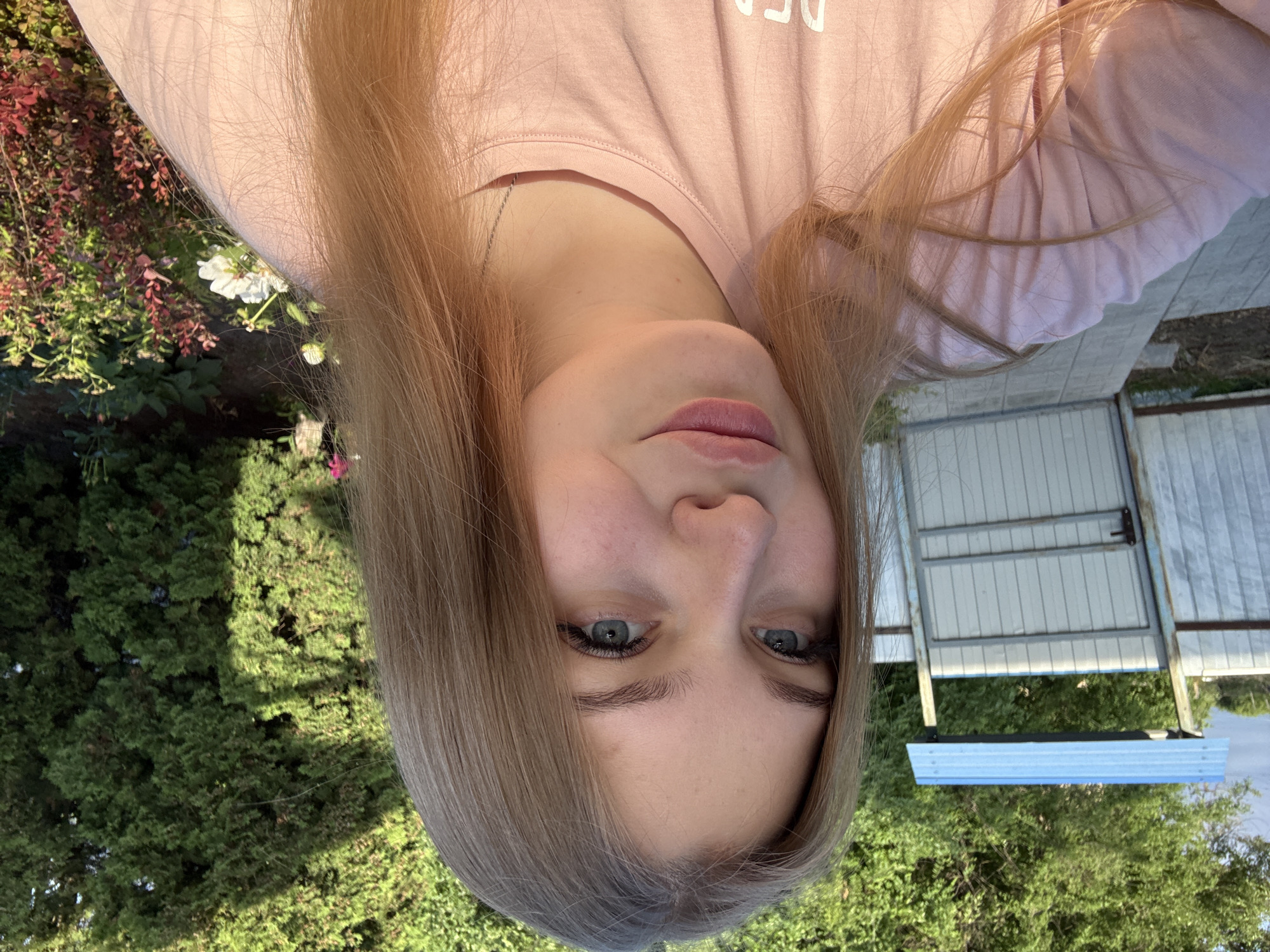 A young woman with blonde hair and blue eyes is taking a selfie outdoors. She is wearing a pink t-shirt and a silver necklace. The background shows a fence, a gate, and lush green trees. The scene appears to be in a residential area or a garden. The image was taken in the city of Sovkhoza "Razdol'e", Russia.