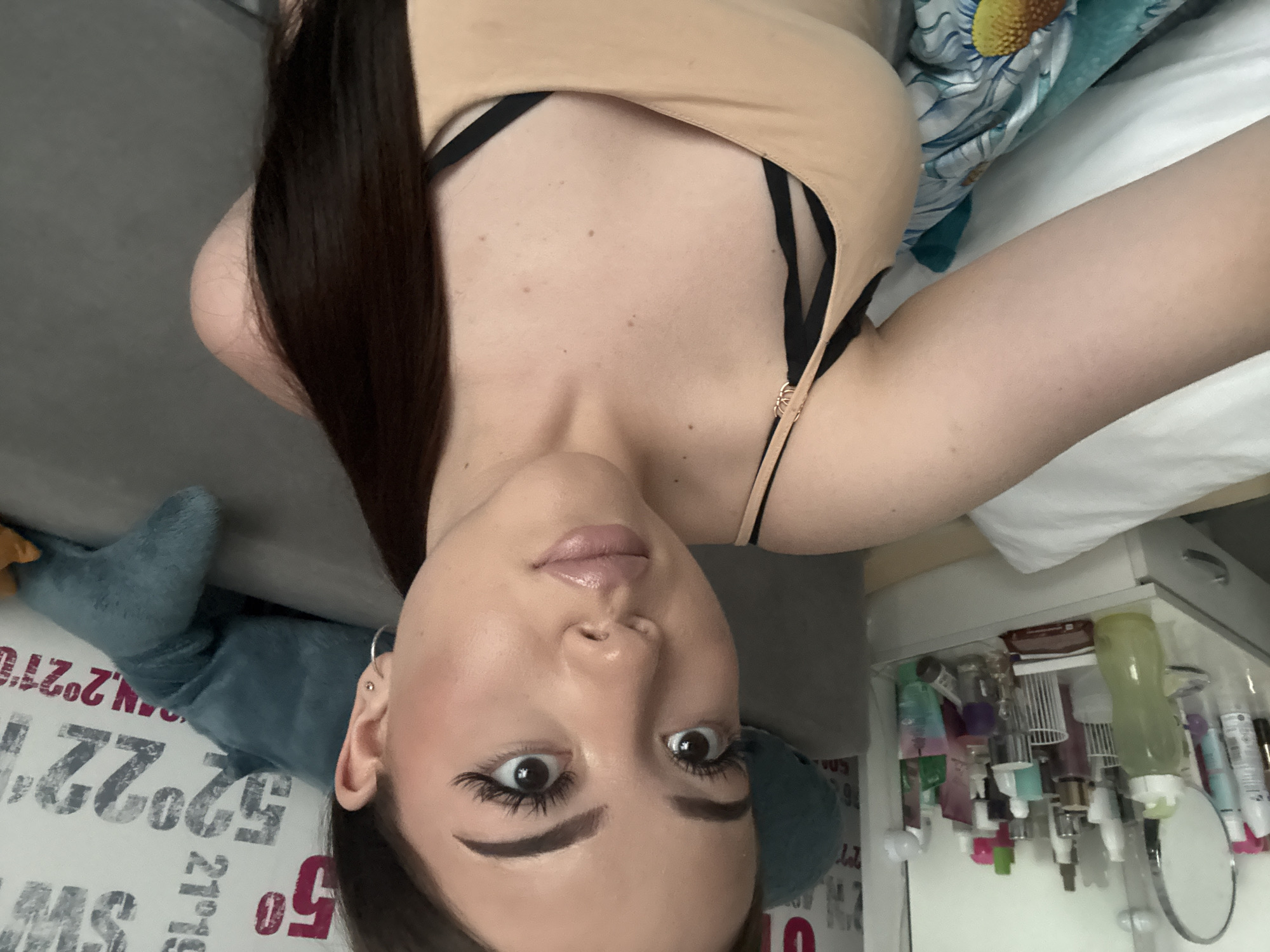 A young woman is reclining in an indoor environment, likely a bedroom. She is wearing a nude-colored top with black straps. The woman has long, dark hair, fair skin, and prominent eyebrows and eyelashes. Her gaze is directed towards the camera.
Behind her, there is a soft, grey-colored surface that could be a couch. A stuffed blue plush toy resembling a shark is also visible. The background also contains a bed covered with a white pillow and patterned blue-green sheet. Beside the bed, there is a cluttered white shelf or drawer filled with cosmetics and personal care items.
The image does not offer any detail related to Naberezhnye Chelny, Russia.