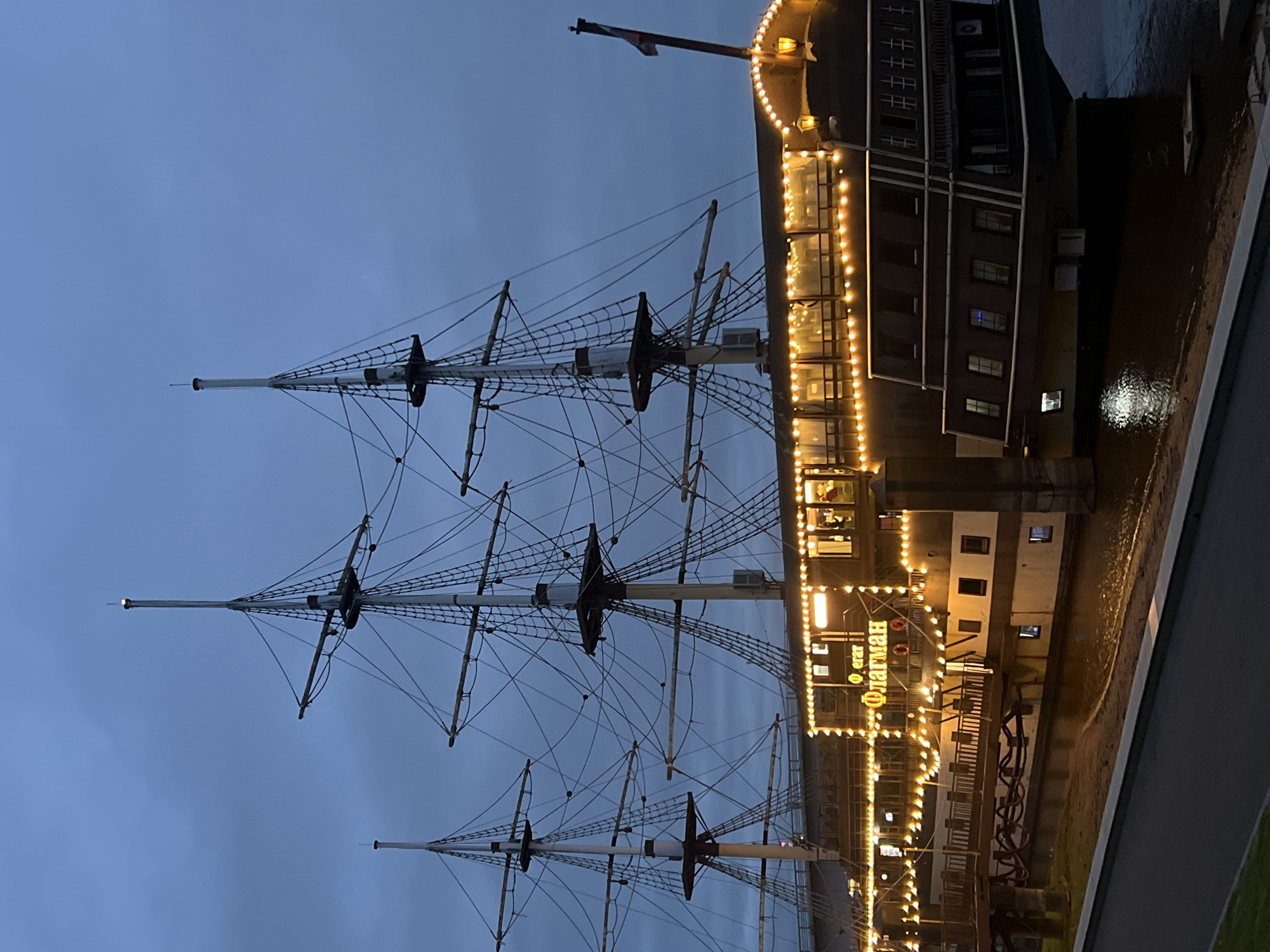 The image shows a tall ship, appearing to be a restaurant or museum, docked at the embankment of the city of Veliky Novgorod, Russia. The ship has three masts and is adorned with numerous string lights that illuminate its decks and signage. The signage on the ship's side reads "Флагман" in Cyrillic, which translates to "Flagship." The sky is overcast and dark, suggesting it is evening or twilight. The ship is moored in a body of water, likely a river or canal, with reflections of the lights visible on the surface. A wooden walkway connects the ship to the shore. The embankment is visible in the foreground, with a paved area and some greenery. There are no people clearly visible in the image.
