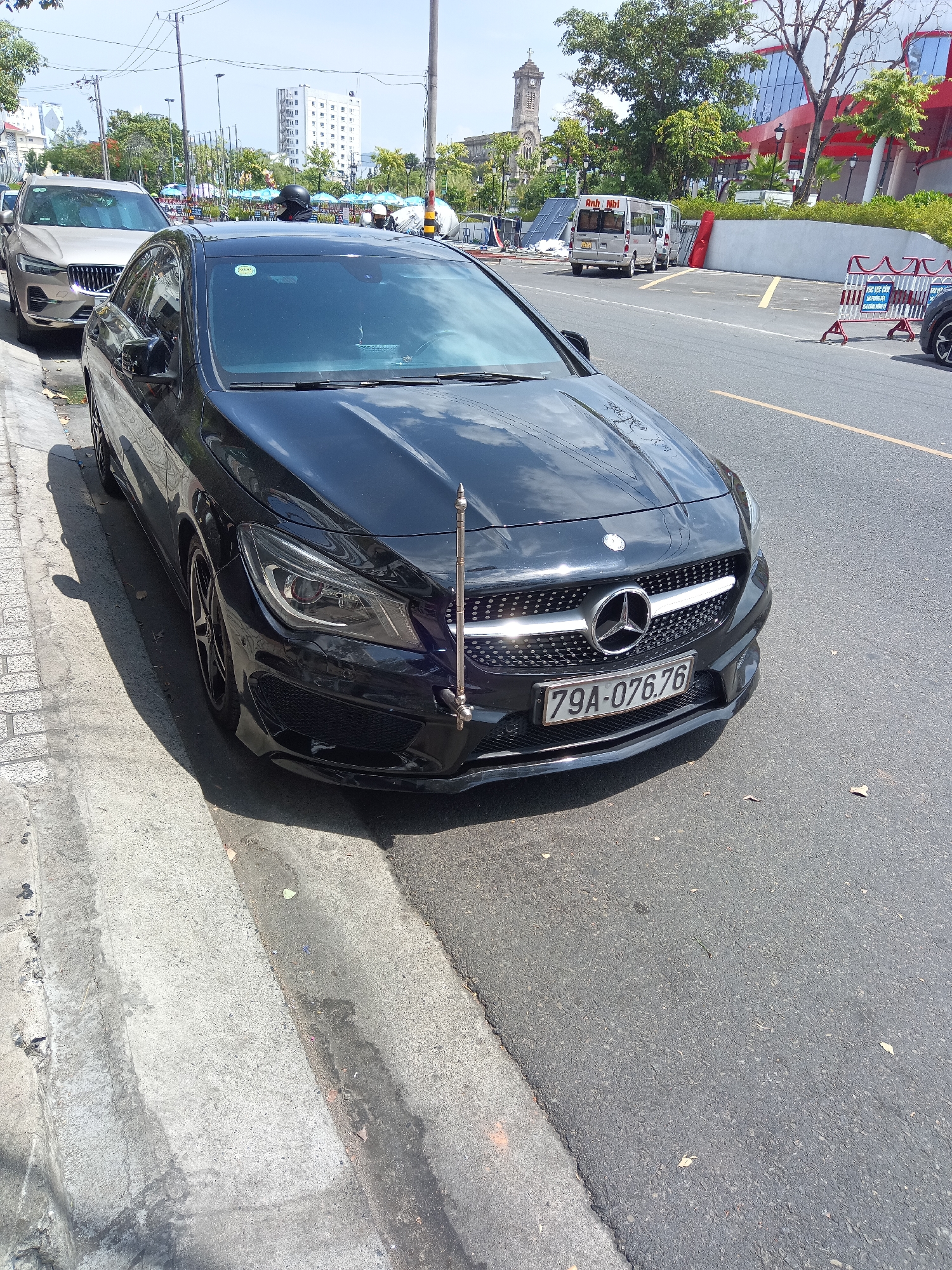 In Nha Trang, Vietnam, a black Mercedes-Benz CLA-Class sedan is parked on the right side of a paved road, its front tires partially on the curb and sidewalk. The car's license plate reads "79A-076.76". In the background, a light-colored SUV is parked to the left of the Mercedes. Further back, buildings, trees, and overhead power lines are visible, along with a street with barriers and a white van. The sky is clear and blue.