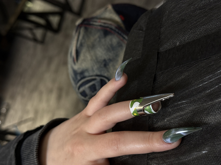 A close-up, eye-level indoor shot captures the left hand of an individual, located in Moscow, Russia. The hand, exhibiting fair skin, emerges from a dark grey ribbed sleeve. The visible fingers (index, middle, ring, and pinky) are adorned with long, sharply pointed artificial nails featuring a translucent green-grey marbled or cat-eye effect design. A metallic silver C-curve pinching clip is affixed to the middle finger, positioned over a flexible nail form. The nail form displays a green and white pattern, including a stylized "M" logo and other graphic elements suggestive of commercial branding. The hand rests upon dark, textured material, possibly leather or faux leather clothing. The background is blurred, revealing indistinct shapes of blue denim fabric and dark furniture or flooring, consistent with an indoor commercial establishment.