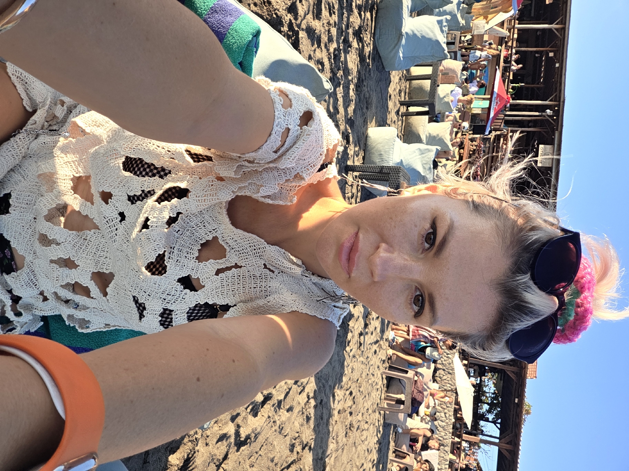 The image depicts a woman in a white lace top, possibly on a beach in North Kuta, Indonesia, based on the presence of sand, beach chairs, and a wooden structure resembling a beachside establishment. The woman is wearing sunglasses perched on top of her head and an orange watch. The environment consists of sand, sun loungers occupied by people, and a partially visible wooden building with people under it. The overall scene suggests a sunny day at a beach resort.