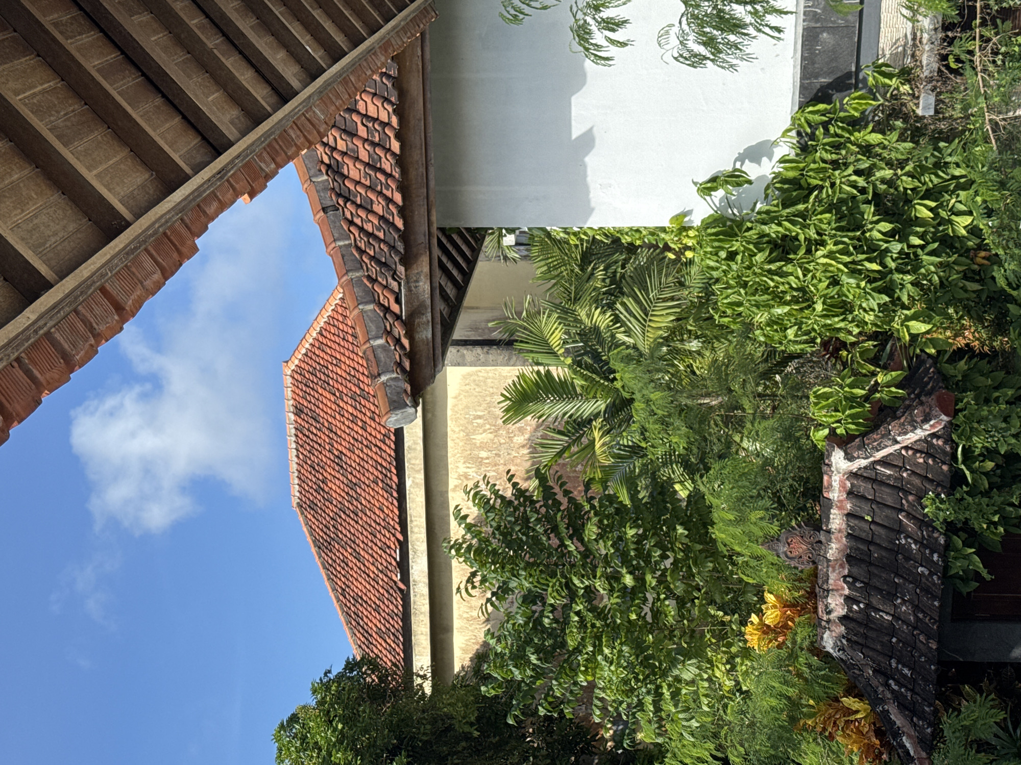 An eye-level, low-angle, outdoor shot reveals a medley of architectural elements and lush vegetation in Kuta Selatan, Indonesia. The frame is dominated by sections of tiled roofs, one a warm red-brown, and another a darker, almost black hue, suggestive of age or a different material. A wooden-slatted overhang adds depth and obscures the view of the sky on the left.
The sky is a clear, bright blue with a smattering of white clouds. Adjacent to the red-brown roof is a beige-colored wall. The greenery is dense and vibrant, with a variety of foliage including palm trees. The composition implies a view from within a complex of buildings or a garden. The light suggests a sunny day, with shadows cast by the plants on the wall.