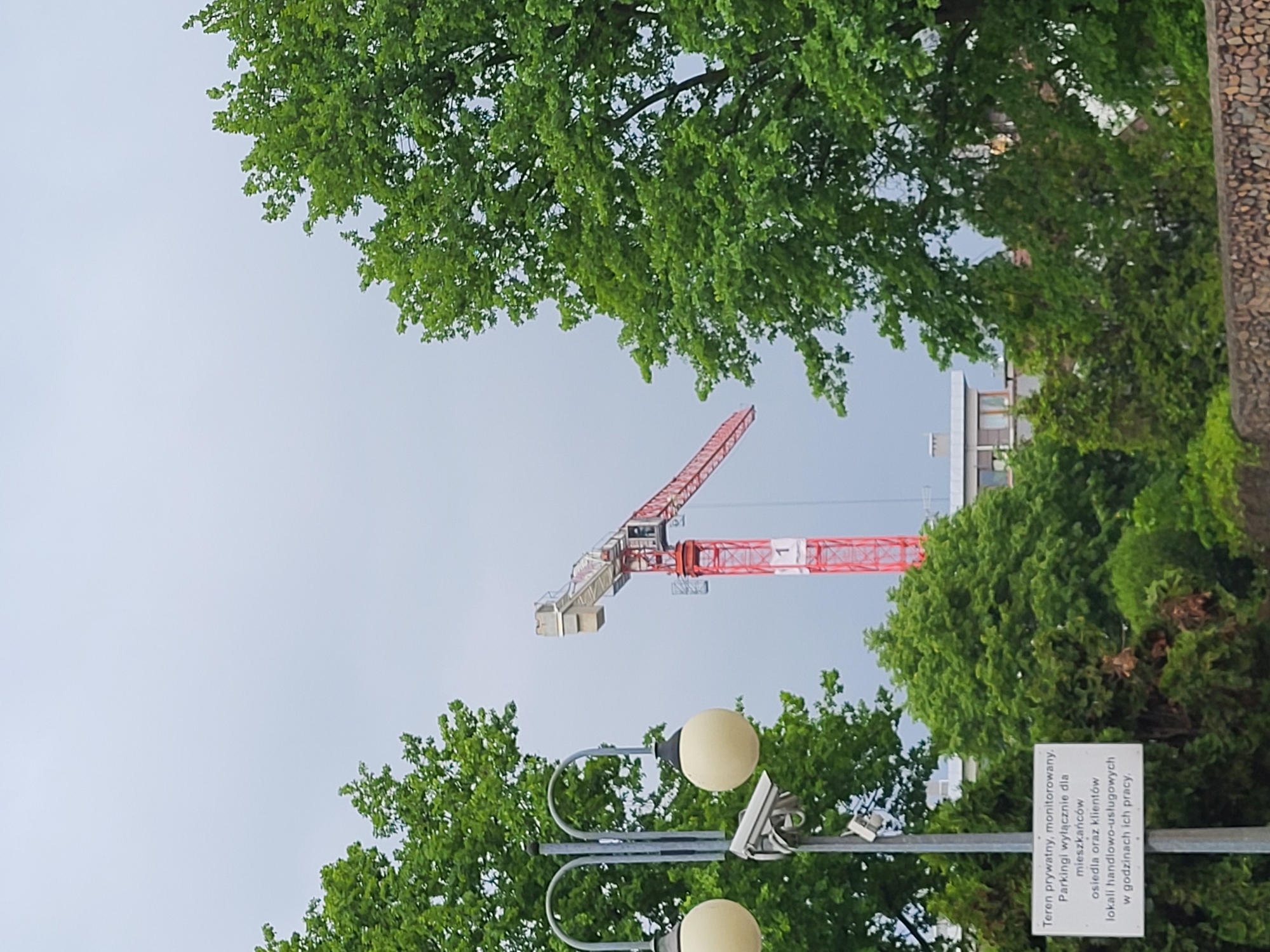 The image shows a crane with a red arm extending into the sky. The crane is surrounded by green trees and foliage, partially obscuring it. In the lower part of the image, there is a street lamp with two round, white lampshades and a security camera mounted on the pole. A white sign with black text in a foreign language is also visible near the street lamp. A building can be glimpsed in the background behind the trees, but its details are not clear. The sky above is overcast.