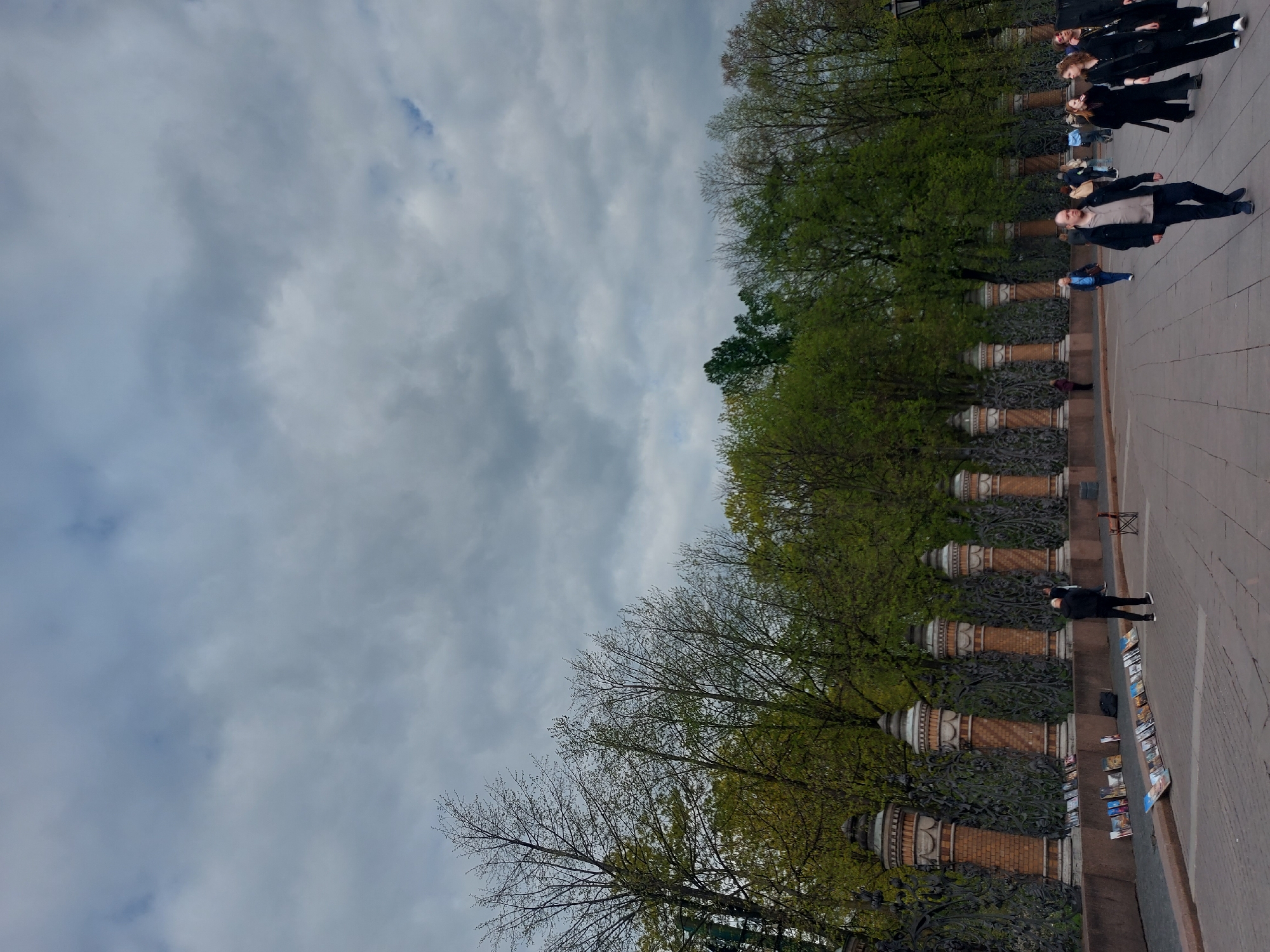 The image shows a street in Saint Petersburg, Russia. On the right side of the image, several people are walking along a paved walkway. A row of trees and ornate brick and iron fence runs along the left side of the walkway. There are trees, some with leaves and others without, behind the fence. On the right side of the walkway, a person is standing near a display of printed materials. The sky is cloudy.