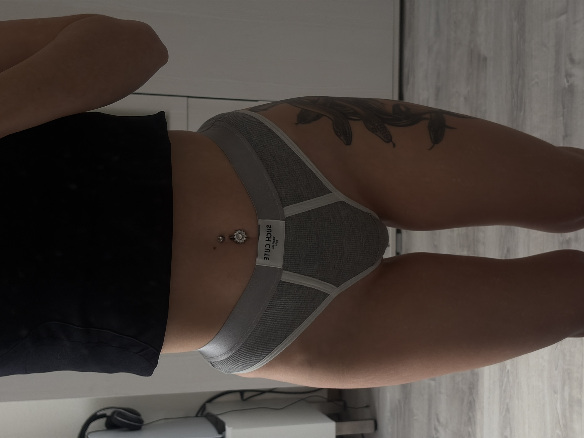 A woman's torso and legs are depicted from a high angle perspective. She is wearing gray underwear with a white band that reads "SUCH CUTE" in reverse. A black top covers her upper torso. The woman has a navel piercing and a tattoo on her right thigh. The background includes a wooden floor and a white surface with electronic devices, possibly a charger and headphones. The location is unspecified and there is no indication of Saint Petersburg, Russia.