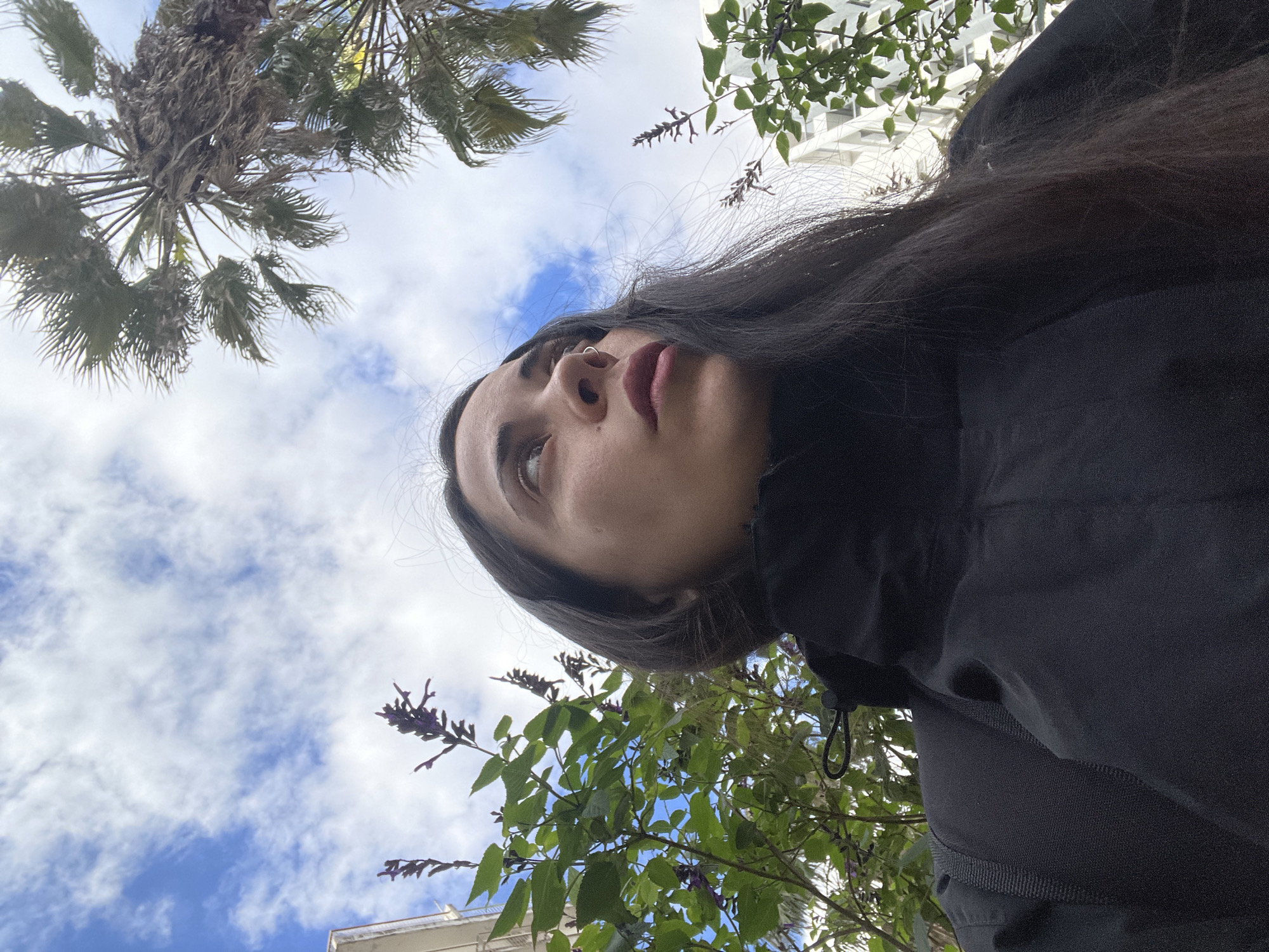 The image, originally rotated 90 degrees clockwise, depicts a young person looking upwards at the sky and surrounding environment from a low-angle perspective. In the lower right foreground, a person, likely female, is shown from the chest upwards. She has long, dark hair and is wearing a dark, high-collared jacket or top. A small, circular nose piercing is visible on her left nostril, and her lips are painted a muted reddish-pink. Her gaze is directed upwards and slightly to the left, conveying a thoughtful or observant expression.
The sky occupies the majority of the upper frame, displaying patches of bright blue with scattered white, fluffy clouds, indicating daytime. On the upper left, the fronds and base of a palm tree are visible against the sky. On the upper right and closer to the person, are branches with green leaves and small clusters of purple flowers or buds, possibly from a flowering plant like lavender. Parts of light-colored buildings are discernible in the background through the vegetation, suggesting an urban or suburban setting.
The scene captures a candid moment outdoors, with the person seemingly contemplating or observing their surroundings. The weather appears clear and pleasant. Given the location context, this scene is likely set in Buenos Aires, Argentina. No text is visible in the image.