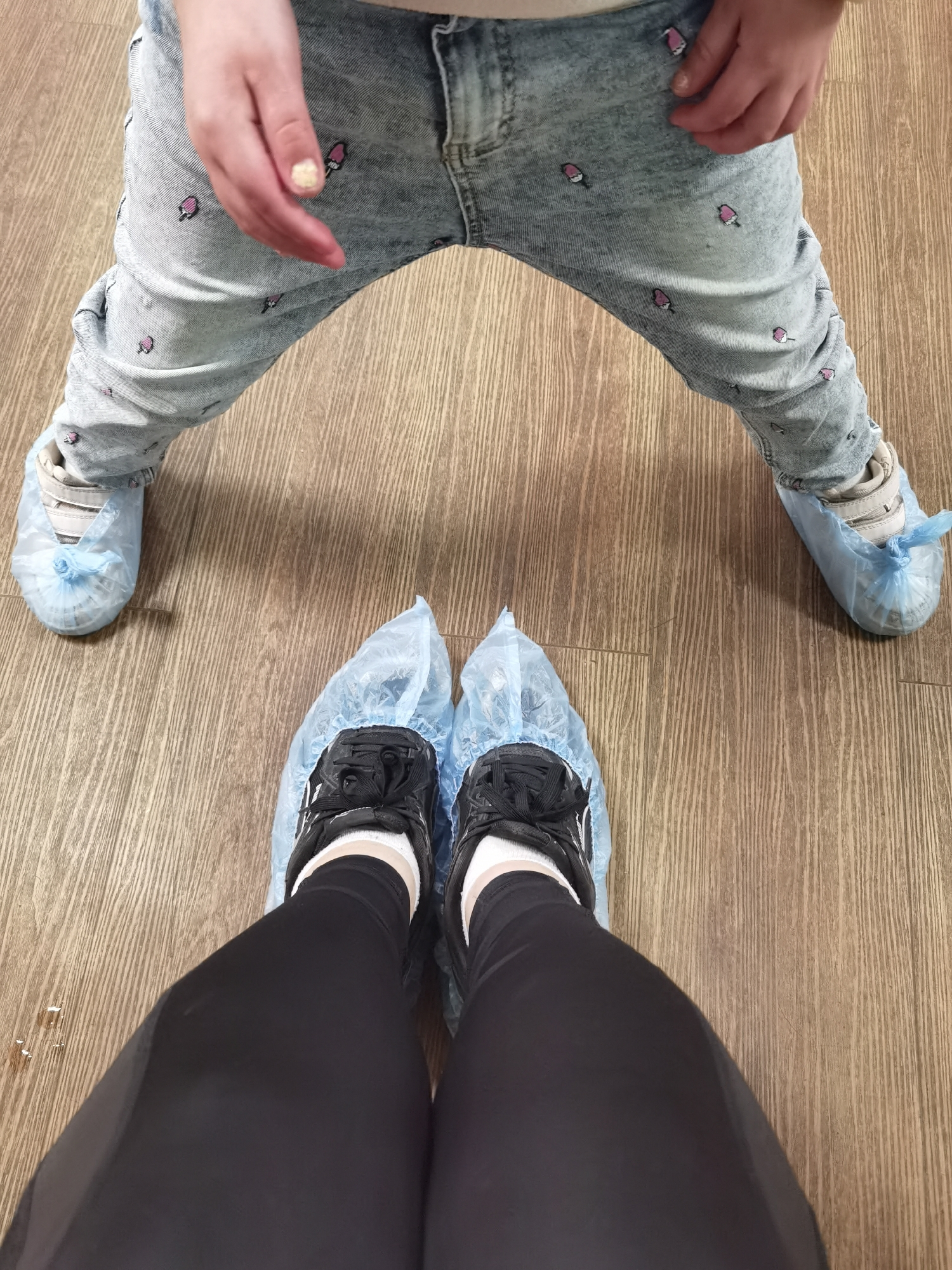 An indoor scene in Izhevsk, Russia, depicts the lower extremities of two individuals, an adult and a child, viewed from a high-angle perspective. Both individuals are wearing light blue disposable shoe covers over their footwear. The flooring is a light to medium brown wood-look laminate or vinyl, exhibiting a vertical grain pattern. In the foreground, an adult's legs are clad in black leggings, white socks, and black athletic shoes. In the background, a child wears light blue denim jeans adorned with multiple small pink popsicle patterns. Their shoes, light-colored and appearing to be trainers with Velcro fastenings, are also covered. The child's left hand is visible resting on their thigh, with one fingernail painted yellow. The disposable shoe covers indicate entry into a controlled or clean environment.