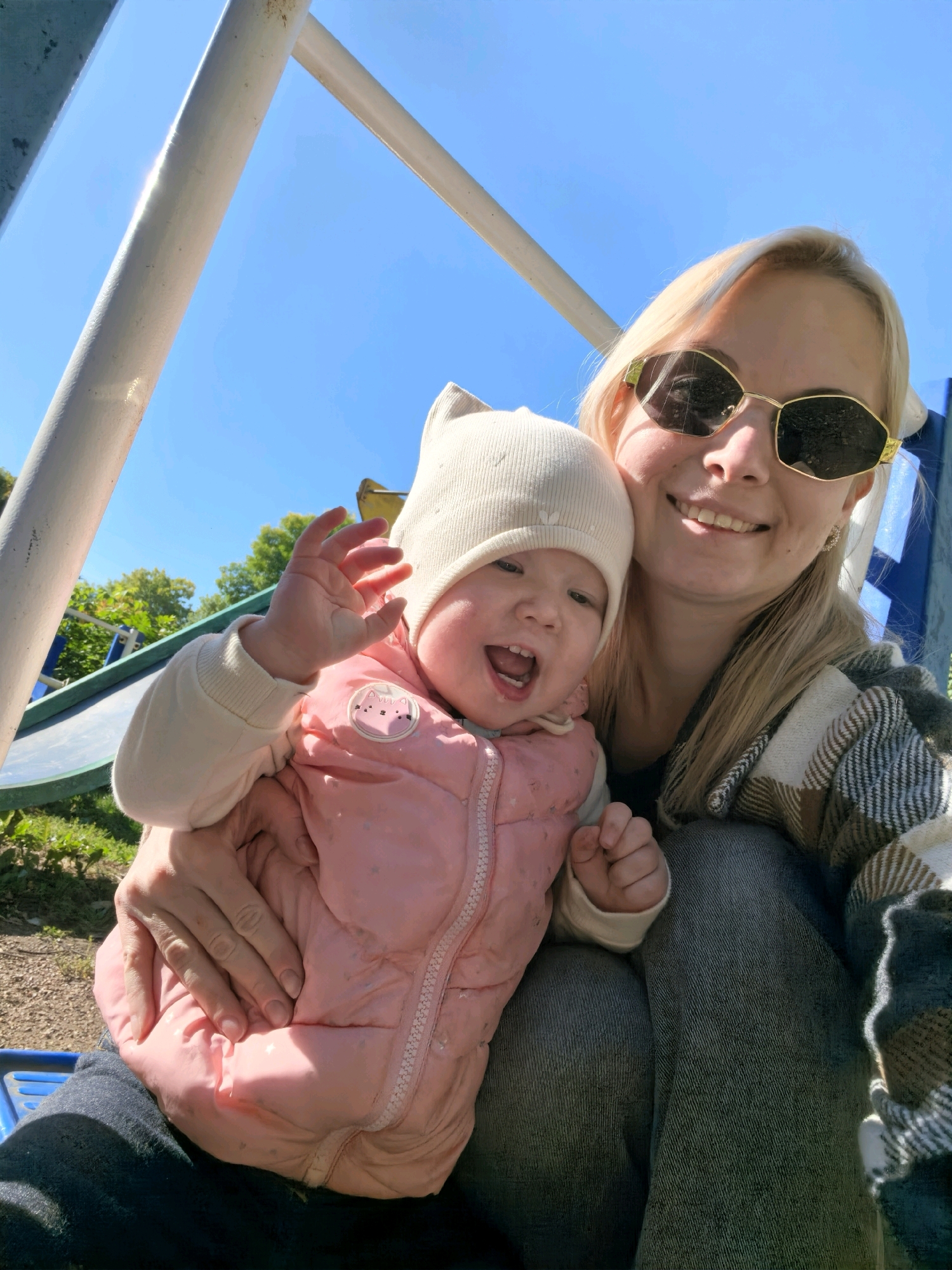 A woman and a baby are sitting on a playground. The woman is wearing sunglasses and a plaid blanket. The baby is wearing a pink puffy jacket and a hat. The sky is blue and there are trees in the background. The image appears to have been taken on a sunny day. There is no information within the image to indicate the location within the city of Sterlitamak, Russia.