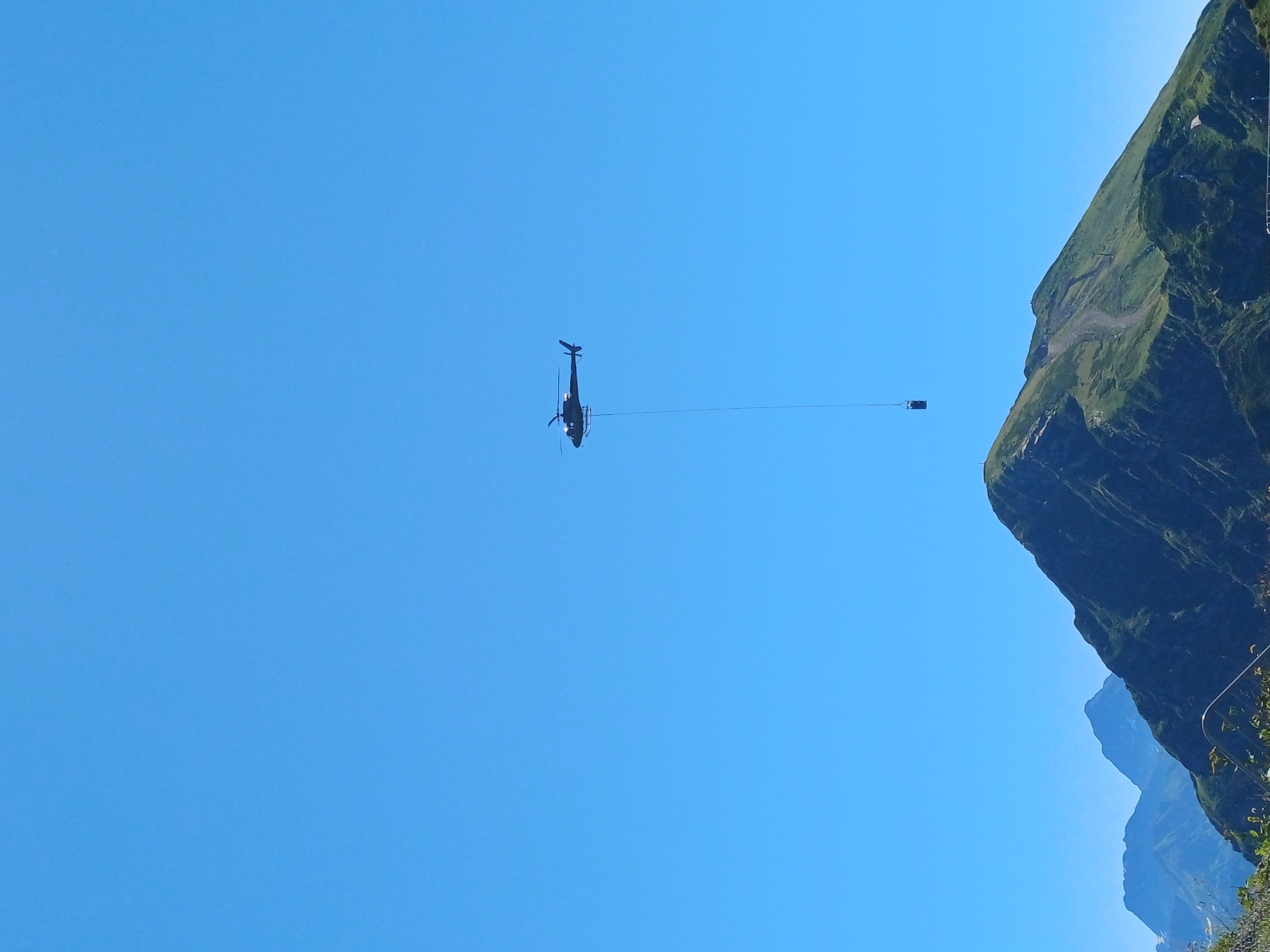 A dark helicopter flies across a clear blue sky above a lush green mountain range. The helicopter is suspended by a long cable, with a small dark rectangular object hanging from the end of the cable. In the foreground, the slopes of the mountains are covered in green vegetation, with a path winding up one of the peaks. In the distance, several other mountain peaks are visible. This scene appears to be in the mountainous region of Estosadok, Russia, known for its ski resorts and outdoor recreation.