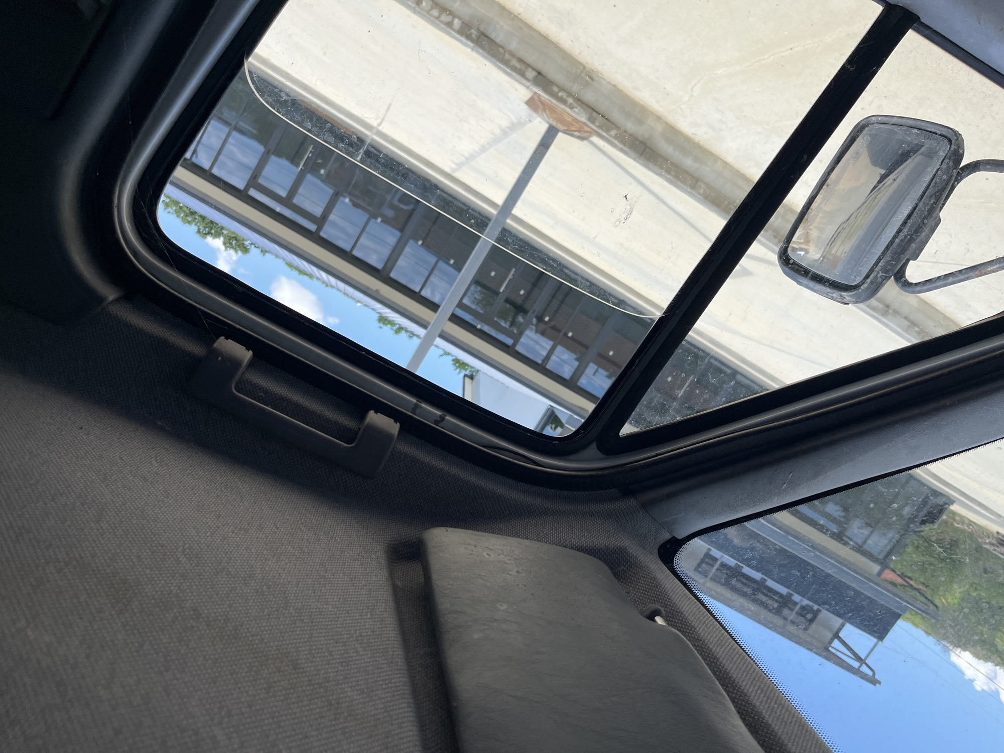 The image provides an interior view from a vehicle, looking out through the windshield and side window. The interior features a gray fabric headliner and a gray sun visor. The side window frame is black and appears to be made of plastic or rubber. A side-view mirror, which is dusty and worn, is visible attached to the exterior of the vehicle.
Through the windows, a modern building with a dark facade and numerous large glass windows is seen. The building appears to be a commercial or institutional structure, possibly with a green roof or vegetation on top. The ground outside the building is a light-colored concrete or asphalt surface, with some signs of wear and tear, including what appears to be a small piece of wood or debris. The sky is blue with some clouds, suggesting it is daytime.
There is no clear indication of the specific location within the city of Prosenik, Bulgaria, based solely on the visual information in the image. The scene depicts a static environment with no people or ongoing actions.