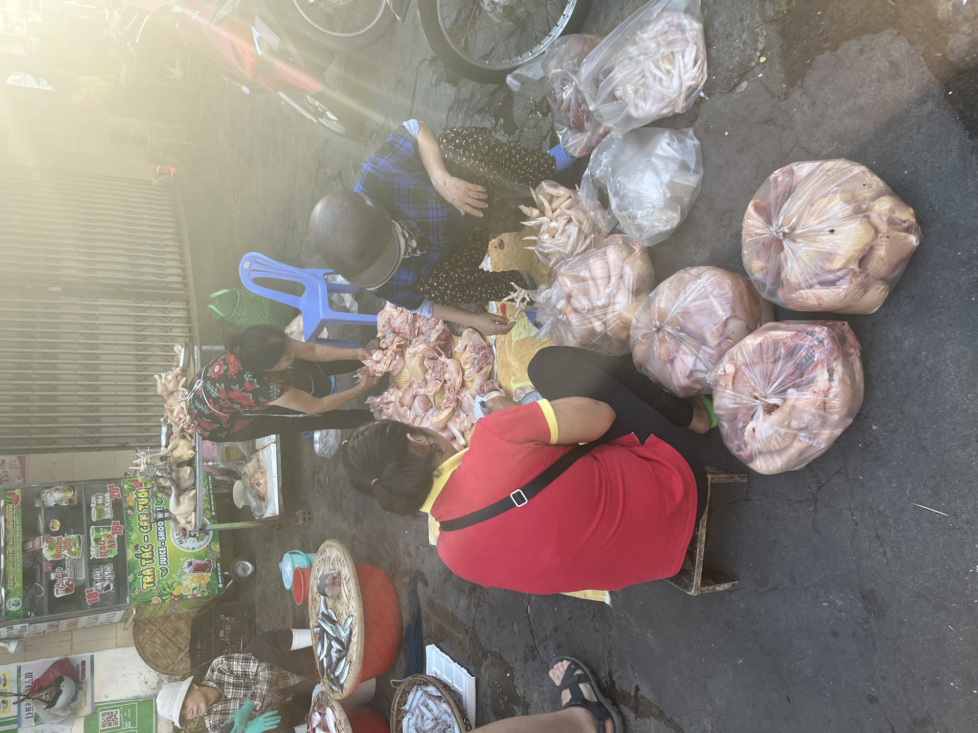 The image depicts an outdoor market scene in Nha Trang, Vietnam, with a strong sun glare. Several individuals are engaged in commerce, primarily focused on fresh produce, including fish and poultry. In the foreground, a woman in a red shirt and black pants sits on a small stool, appearing to be preparing or packaging food items. To her right, another person, wearing a plaid shirt and a helmet, is crouched down, also tending to food products laid out on a tarp. Behind them, a vendor in a patterned apron is seen working near a refrigerated cart displaying beverages and possibly other food items. The ground is asphalt, showing signs of wear and some moisture. Various baskets and plastic bags containing raw chickens and fish are scattered around the selling area. A blue plastic chair and green baskets are also visible. The presence of a motorcycle on the right side suggests this is a roadside market. The overall impression is one of a bustling, everyday transaction taking place in a typical Vietnamese urban setting.