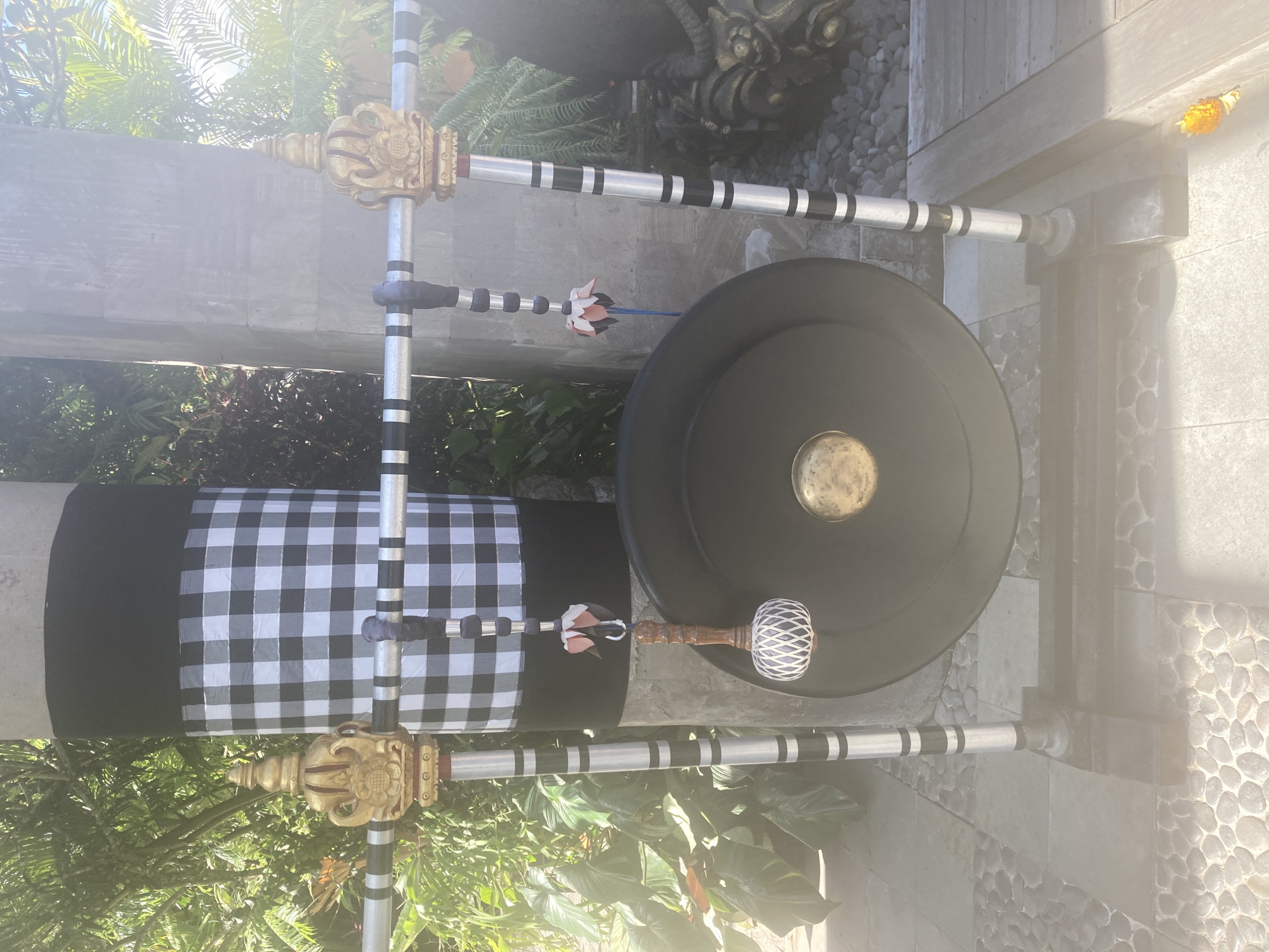 A gong stands outdoors in Unknown, Unknown. The gong is large and black, with a brassy central dome. It rests between two silver poles banded with black stripes. The poles are decorated at the top with golden ornamental fixtures. A black-and-white checkered cloth is draped on the left vertical pole. A mallet with a white-wrapped head rests horizontally in front of the gong. A small flower or ornament is suspended by a string near the gong's upper left. The gong is set against a background that includes lush green foliage and sections of a stone walkway. The overall lighting is bright, casting shadows and creating glare.