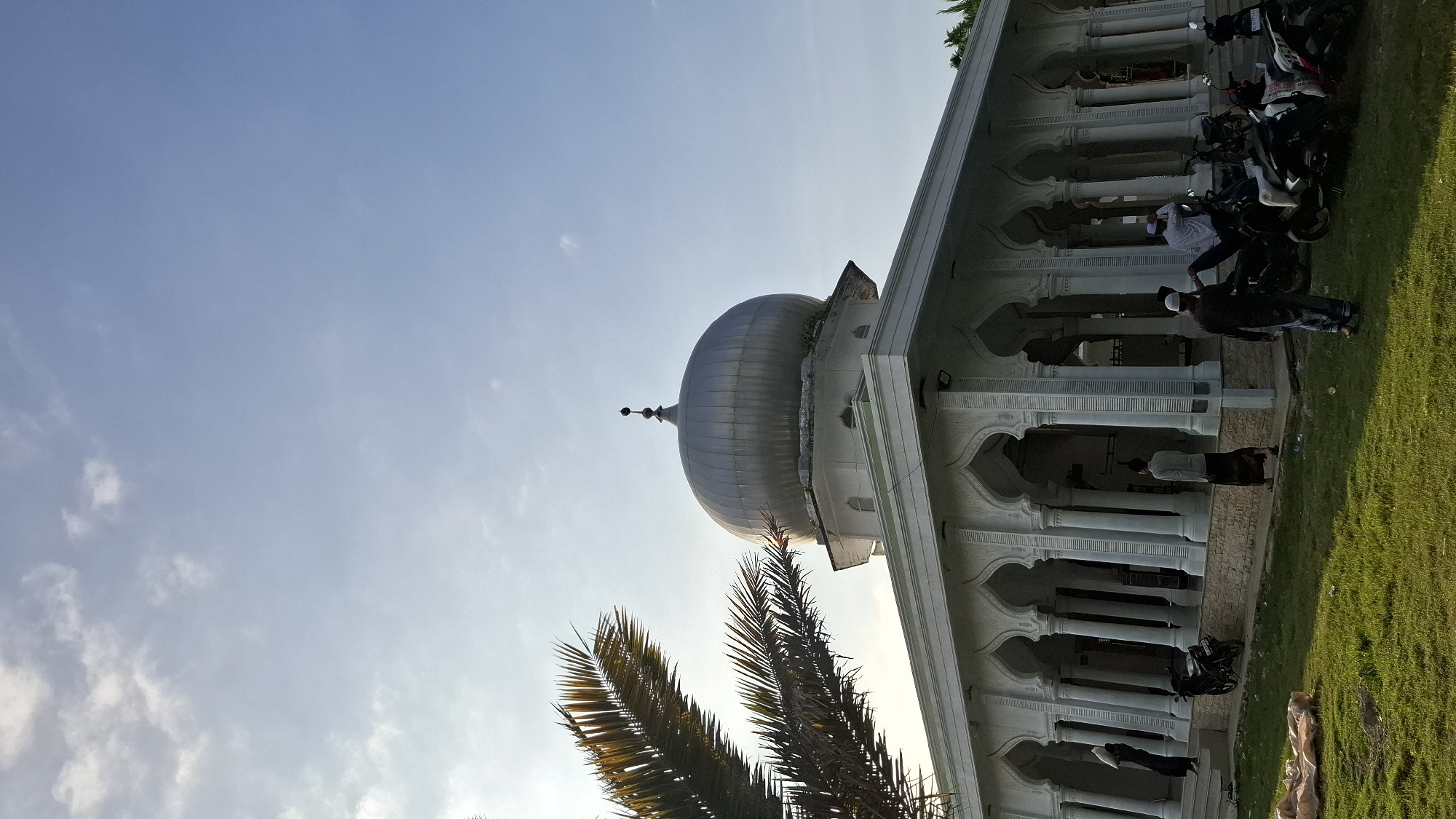 An exterior, low-angle, vertical, medium shot captures a mosque in Grong-grong, Indonesia, under a bright, partly cloudy sky. The mosque features a large, silver dome with a finial, perched atop a white, multi-tiered structure. A colonnaded walkway with arched openings surrounds the main building, supported by white pillars.
In the foreground, a well-maintained green lawn covers the ground. Several motorcycles are parked on the grass in front of the colonnade. A person dressed in dark clothing and a white cap stands facing away from the camera near the parked motorcycles. Another person in a light-colored shirt and dark sarong walks to the left, away from the camera. A woman in black is visible walking up steps to the left of the mosque, carrying a white object.
To the left of the mosque, a large palm frond extends into the frame, partially obscuring the sky and the dome. The overall scene is serene, with a sense of community gathering around the religious site. The lighting suggests it is either early morning or late afternoon, with the sun casting a gentle glow.