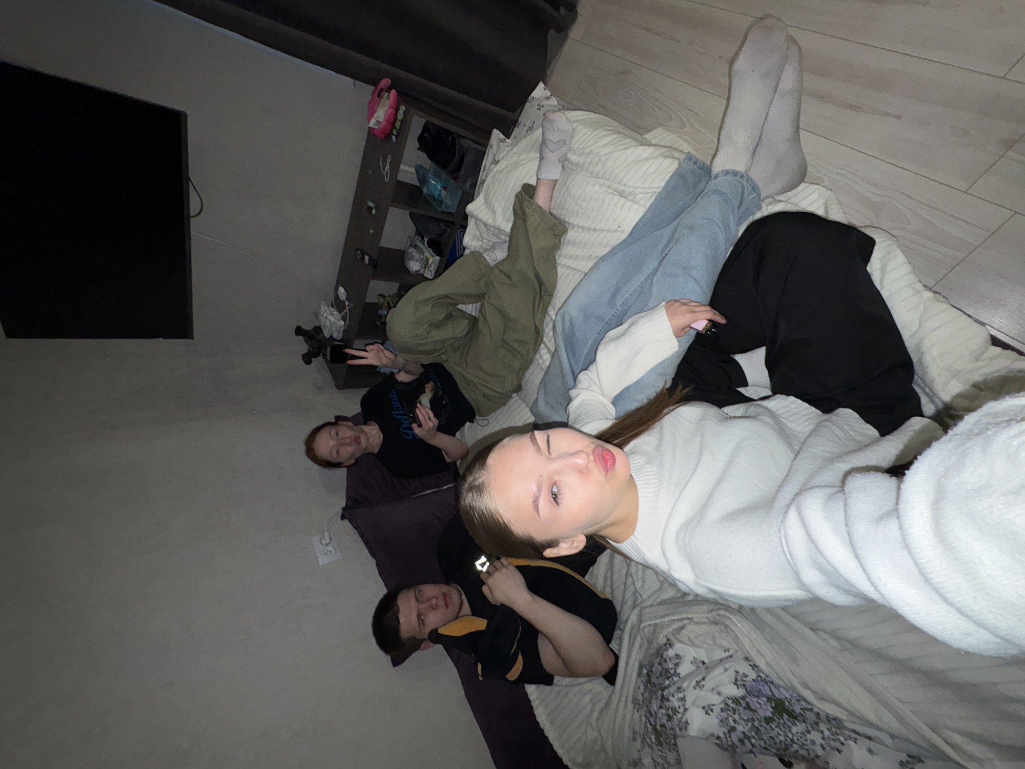 The scene appears to be inside a residence. Four individuals are present, lying on a cushioned surface that could be a bed or large floor cushions. One person wears light blue jeans and grey socks, while another is in olive green pants. A third individual wears a black t-shirt and is making a peace sign with their fingers. The fourth person, closest to the viewpoint, wears a white sweater and seems to be taking the photograph. Other individuals are wearing black clothing. Visible objects include a TV on the wall, a bookshelf with items, and an electrical outlet. The floor is covered with wood-like planks. The environment seems casual and relaxed. It is difficult to confirm the location in Chaikovsky, Russia based solely on the visual information.