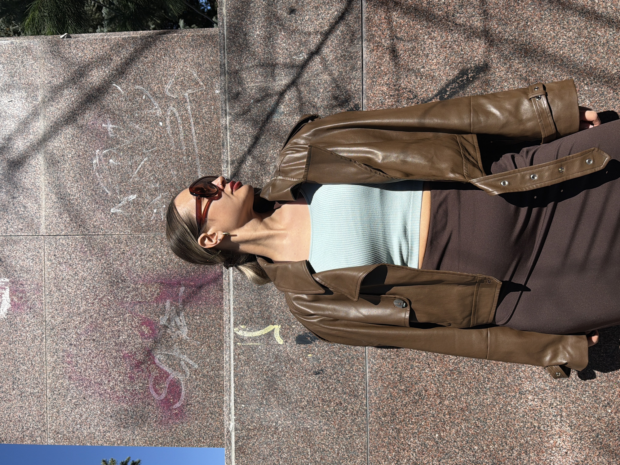 An outdoor, eye-level shot, rotated 90 degrees to the left, features a person, likely female, reclining or lying on a textured granite surface under bright sunlight. The person has light brown hair, pulled back, and is wearing brown sunglasses perched on her head. Red lipstick is visible. She is dressed in a brown, leather-look jacket over a light blue ribbed crop top, paired with a dark brown skirt or trousers. Her head is tilted back, and her eyes are not visible, suggesting she might be looking up at the sky or resting.
The background consists primarily of a mottled granite wall or ground, displaying speckles of brown, pink, and grey. Various graffiti markings are present on the granite, including white scrawls that form indistinct letters, and patches of pink spray paint. One clear white "S" shape is visible, and other white marks resemble letters or symbols. Strong, angular shadows from an unseen object, possibly a tree, fall across the granite surface and parts of the person's body, indicating a sunny day with the sun at an angle. A vibrant blue sky and green foliage from a tree are visible in the lower and upper left corners of the frame, respectively (when viewed upright). The overall scene conveys a casual, urban outdoor setting in Krasnoyarsk, Russia.