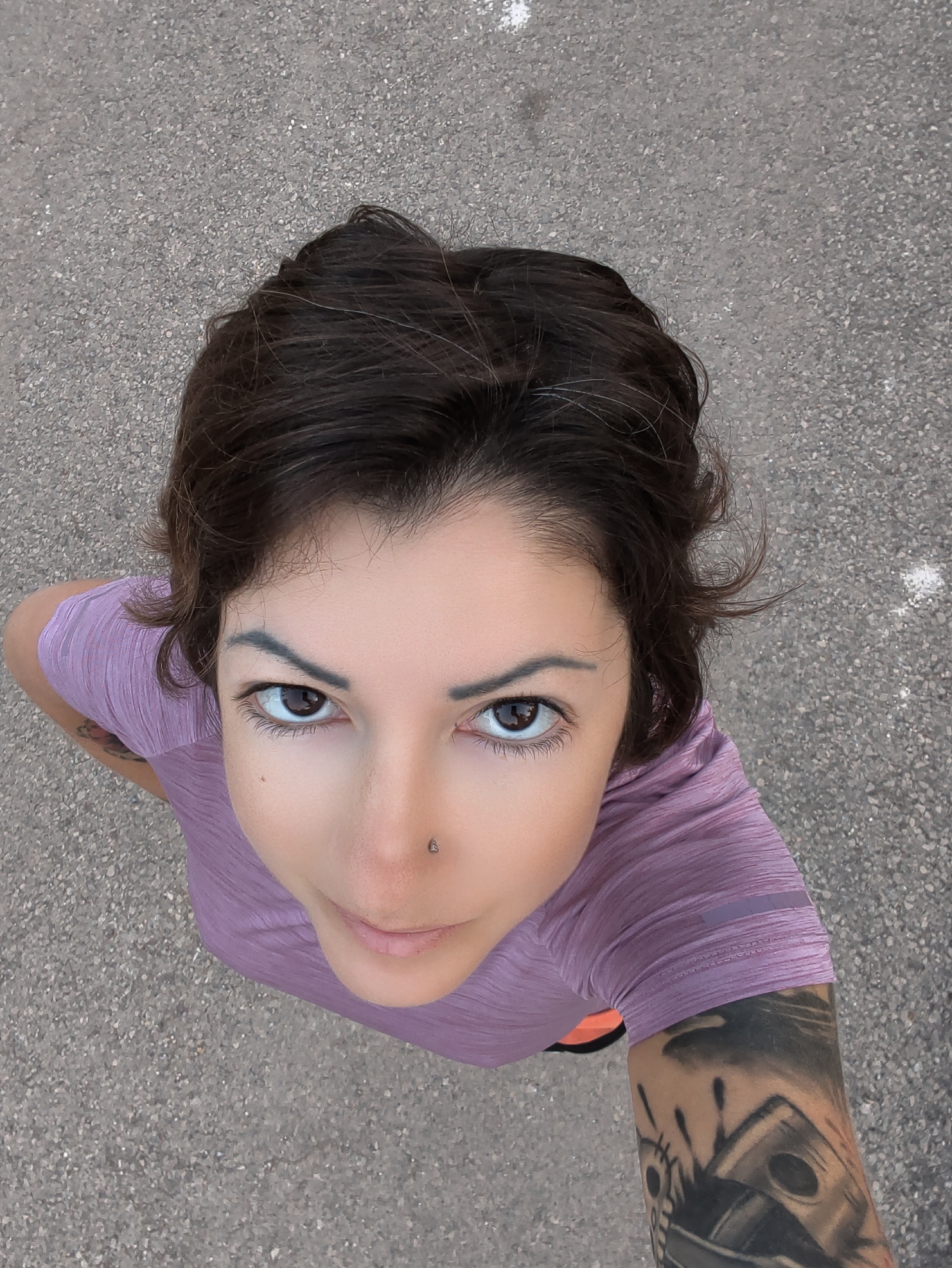 An overhead, close-up, eye-level shot shows a woman from the neck up, looking directly into the camera. She has short, dark brown hair, which is slightly messy, with some strands framing her face. Her skin is fair, and she has dark eyebrows and dark brown eyes. She is wearing a purple t-shirt and has a nose piercing. A tattoo is visible on her left arm. The background is a gray, asphalt-like surface, with a few small white speckles. The overall scene appears to be outdoors, on a paved surface. No specific location in the city of Lachish Regional Council, Israel can be determined from the visual information alone.