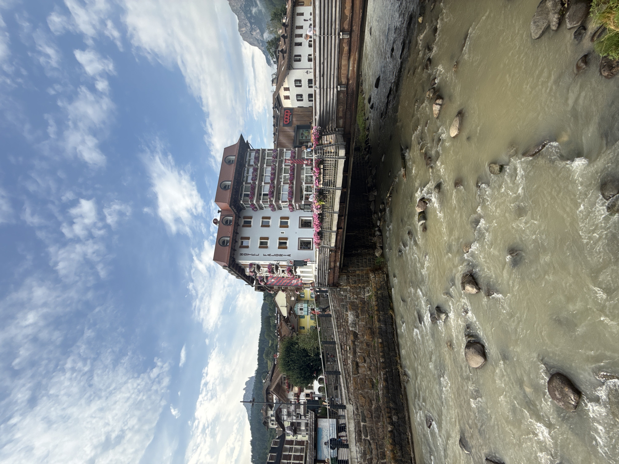 In Moena, Italy, a clear sky with scattered clouds hovers over a bustling scene. A multistory building, identified by the sign "HOTEL FALORIA," stands prominently on the right side of the image. The hotel is adorned with numerous balconies overflowing with pink flowers, and a smaller red sign above the entrance indicates the "Coop" supermarket.
A bridge crosses a wide, rocky river in the foreground, with several large stones visible in the murky, flowing water. People are seen walking across the bridge, which is lined with metal railings and stone walls. The buildings on the left side of the bridge showcase a variety of architectural styles, with colorful facades and traditional roofs. In the background, a majestic mountain range rises against the horizon, its peaks partially shrouded in mist. The overall atmosphere is one of a charming European town with a vibrant natural setting.