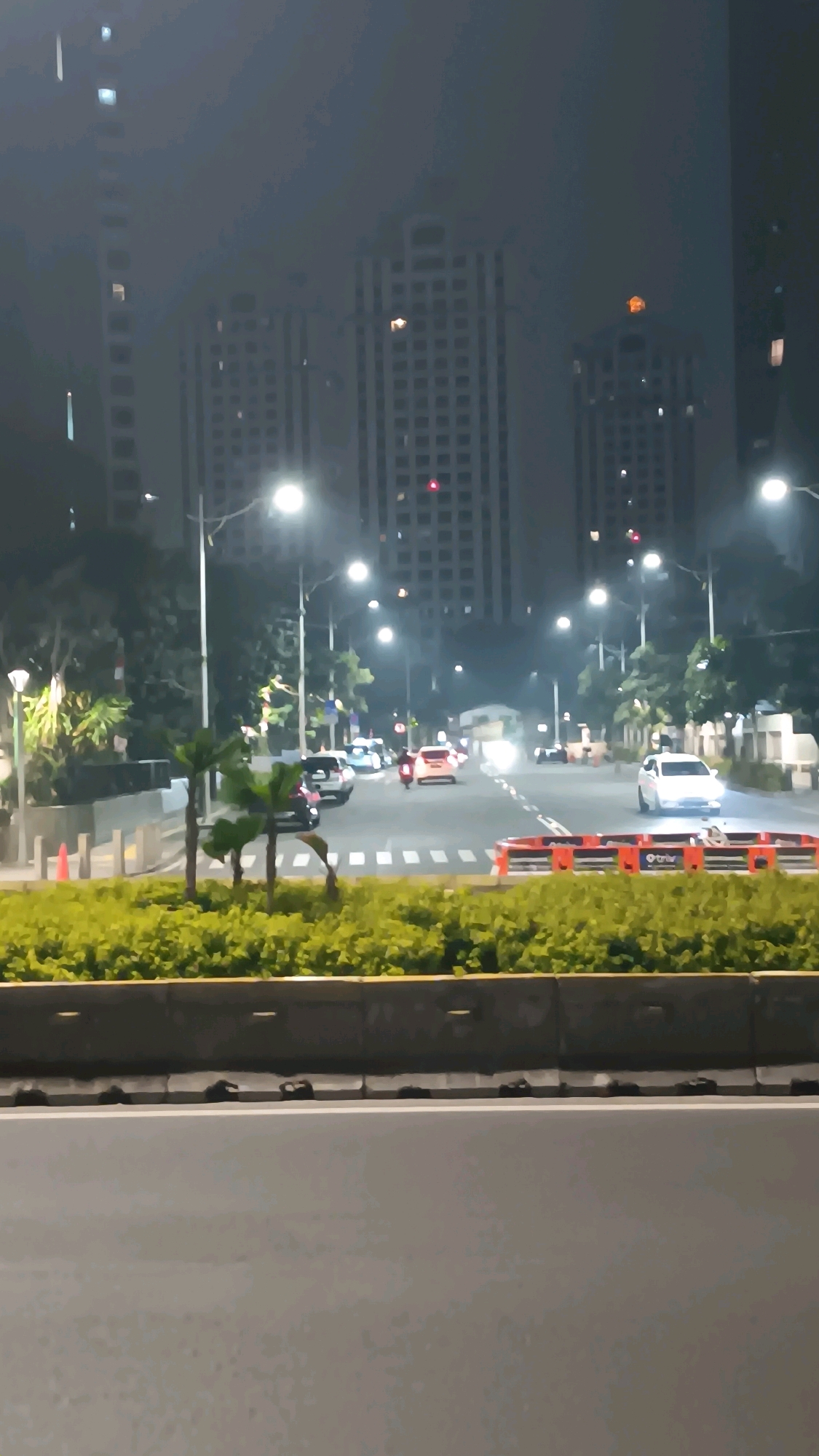 The image depicts a street scene at night in South Jakarta, Indonesia. Tall, multi-story buildings with many illuminated windows dominate the background, suggesting an urban setting. A road with multiple lanes is visible, with several vehicles, including cars and a motorcycle, driving or stopped. Streetlights illuminate the road and surrounding trees. In the foreground, there is a median strip with lush green foliage, separated from the road by a concrete barrier. Orange traffic cones and barriers are also present on the road. The overall atmosphere is dim and hazy, possibly due to weather conditions or light pollution.