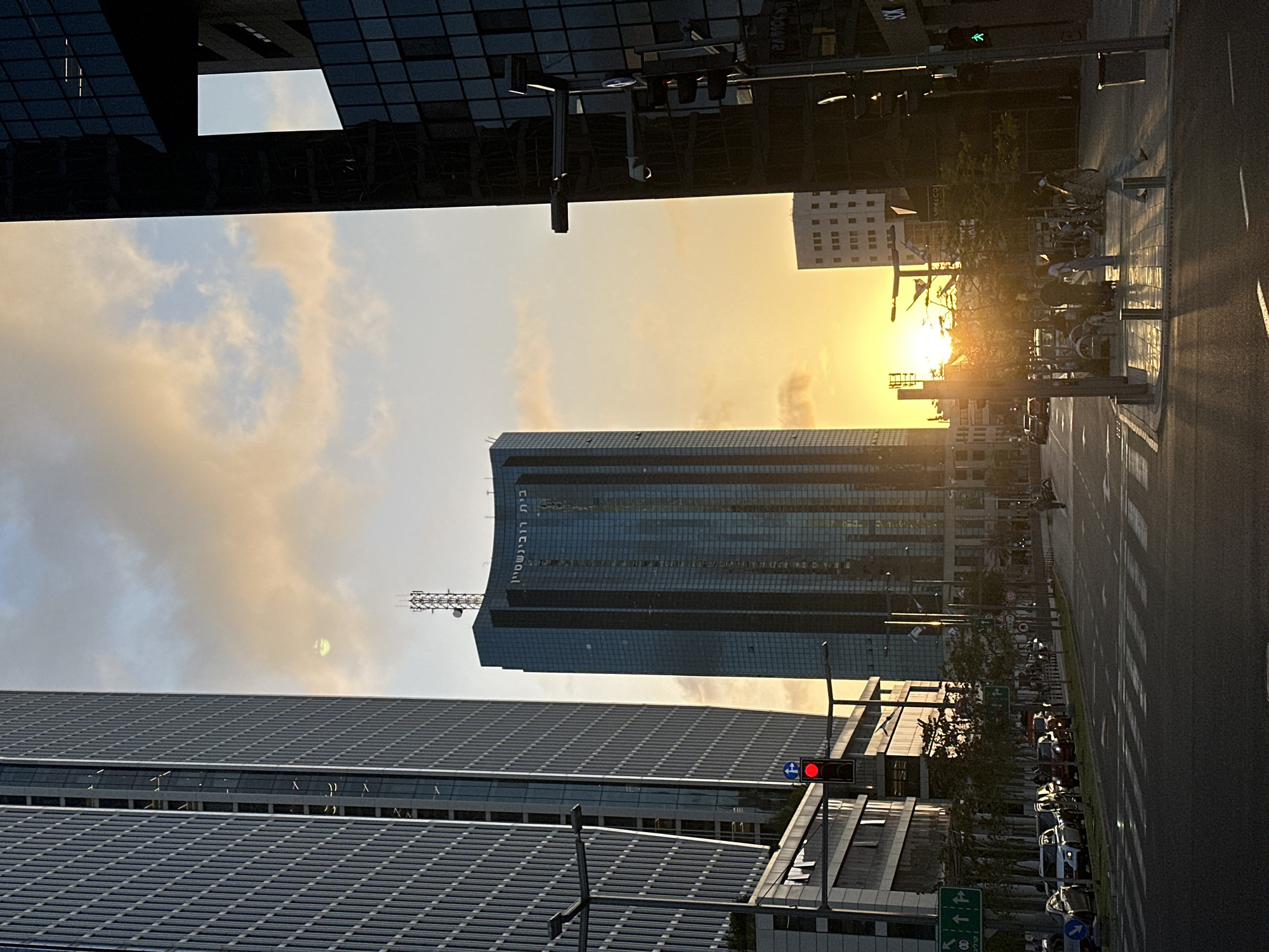 This image captures a vibrant urban street scene in Tel-Aviv, Israel, during sunset or late afternoon, characterized by dramatic lighting and modern architecture. The sky transitions from a brilliant, low-hanging sun on the right, casting warm orange and yellow hues, to lighter blue and scattered clouds higher up. The sun's intensity creates significant glare and lens flare across the scene.
Dominating the central right is a tall, glass-clad skyscraper, reflecting the ambient light and sky. Vertically written Hebrew text, "בית רובינשטיין" (Beit Rubinstein - Rubinstein House), is visible on its side, and an antenna array tops its roof. Another large building with a textured, grey facade occupies the left foreground, while dark, reflective structures frame the upper portion of the scene. In the background on the far right, a lighter-colored building is partially visible.
At street level, a multi-lane asphalt road stretches across the bottom-right, marked with lane lines and long, distinct shadows. A tree-lined sidewalk or median separates the road from the main buildings. A red traffic light and a blue circular 'turn right' sign with a white arrow are visible on the left. Further down the street, a green pedestrian traffic light and a green directional sign can be seen. Several cars are parked along the street, and faint silhouettes of pedestrians are discernible on the sunlit sidewalk. The scene portrays a bustling city environment as the day concludes, with the low sun emphasizing the architectural forms and street activity.