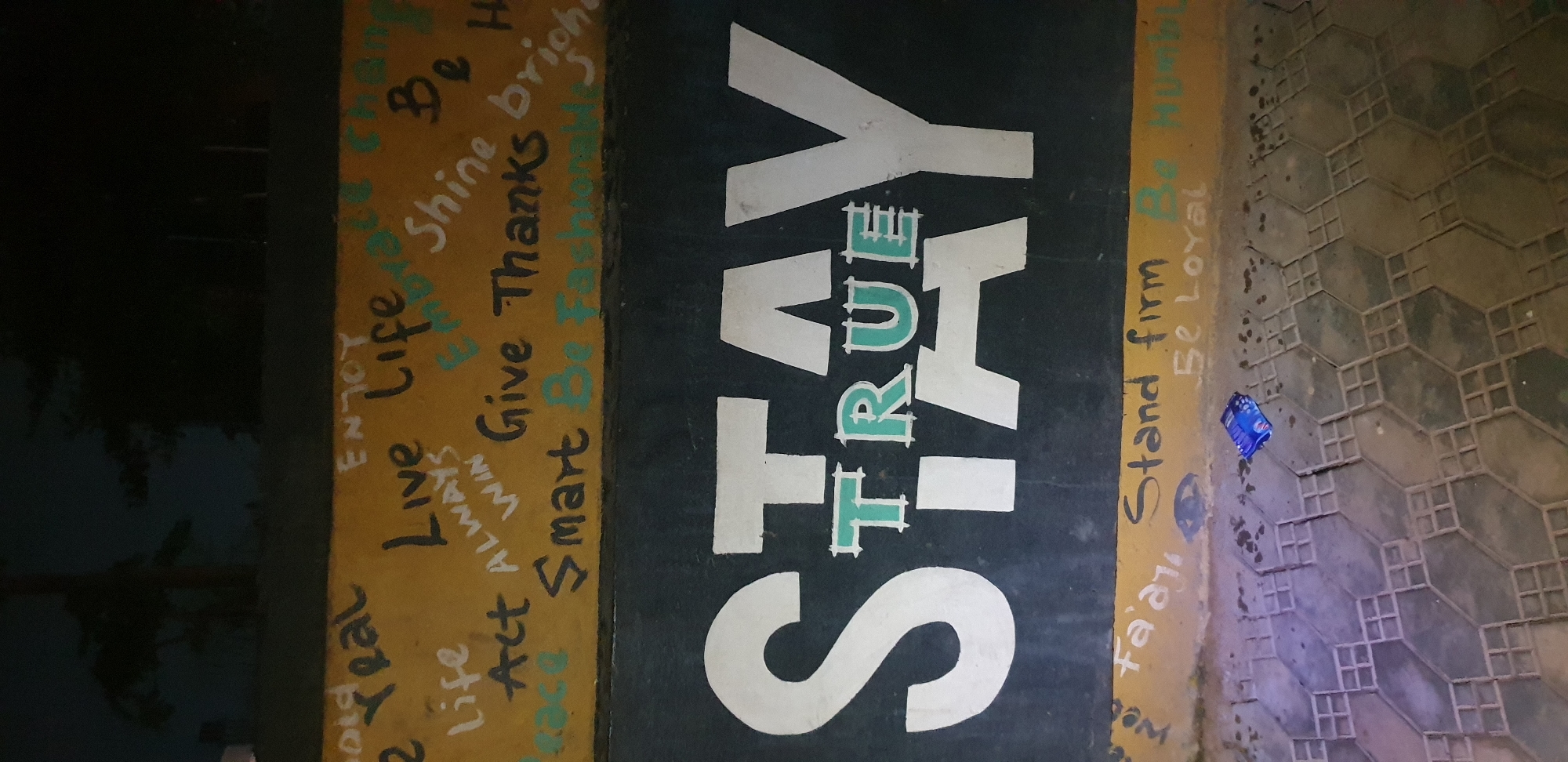 The image shows a wall in Narayi, Nigeria, with graffiti. The graffiti includes the words "STAY TRUE" in large white letters on a black background, and smaller phrases like "Be Real," "Enjoy Life," "Act," "Always Win," "Give Thanks," "Smart," "Embrace change," "Shine bright," "Stand firm," "Be Loyal," and "Be Humble" written in various colors on a yellow background above the large text. The wall appears to be part of an outdoor space, possibly a street or a pathway, as indicated by the tiled ground in the foreground. There is a discarded blue wrapper on the ground. The lighting suggests it is nighttime or a dimly lit area. There are no people visible in the image.