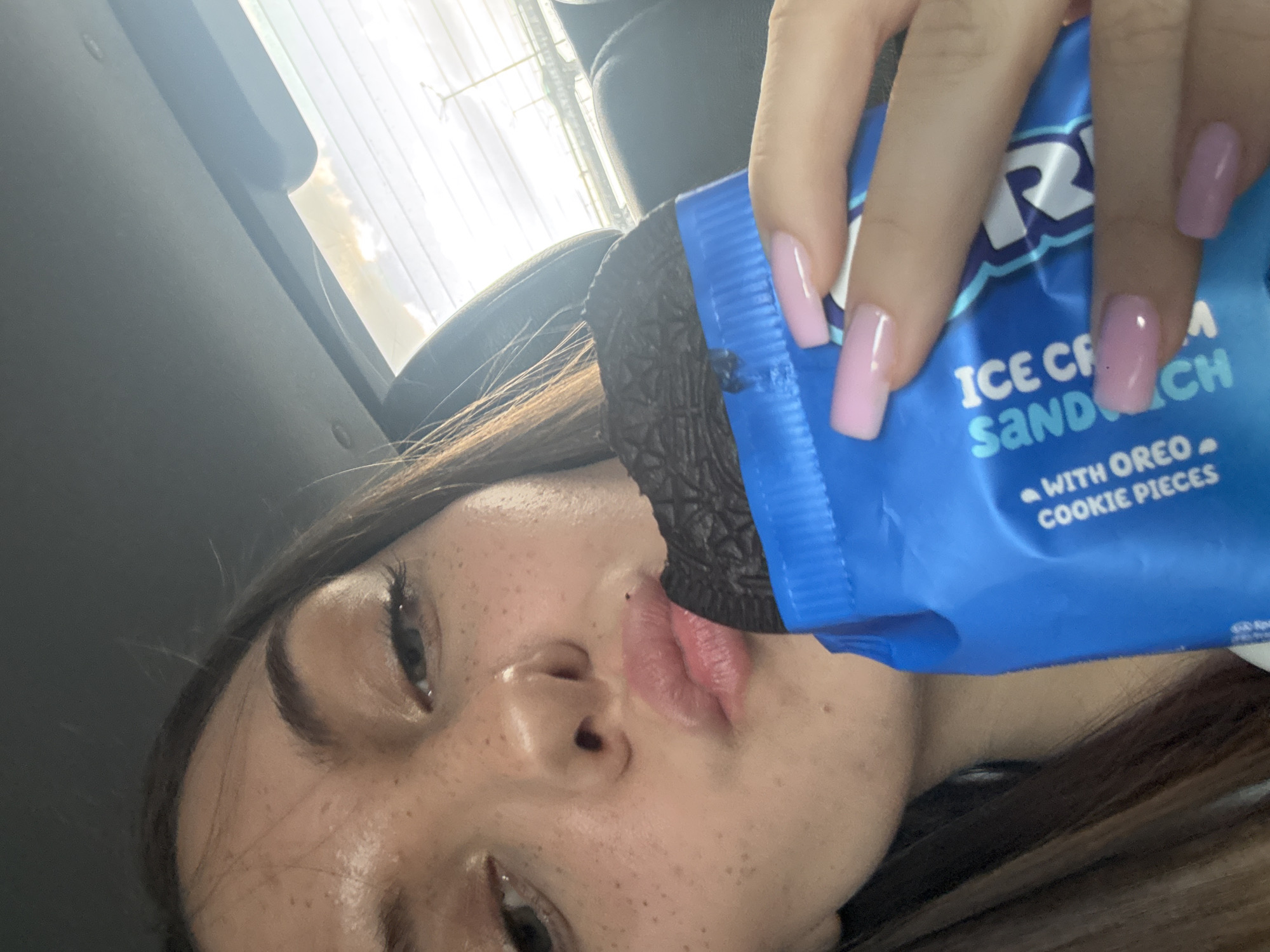 A close-up, high-angle, and slightly tilted photograph captures a person eating an Oreo Ice Cream Sandwich inside what appears to be a car, likely in Sofia, Bulgaria. The central subject is a young woman, partially seen from her eyes down to her chin, with her face slightly turned. She has prominent freckles across her nose and cheeks, and her long, dark brown hair frames her face.
She is holding a bright blue Oreo brand wrapper with her right hand, bringing the ice cream sandwich to her mouth. Her hand displays well-manicured, rectangular nails painted in a light, translucent pink. The dark black Oreo cookie portion of the ice cream sandwich is visible as she takes a bite, pressed against her lips.
Text on the blue wrapper clearly shows "ICE CREAM SANDWICH" and "WITH OREO COOKIE PIECES," along with a partially visible "OREO" logo (the "R" and "O" are discernible).
The background suggests an indoor setting, with a dark interior surface (possibly a car headliner or dashboard) visible in the upper left, and a car seat or headrest on the right. Through a window in the upper background, bright daylight and a hazy sky are visible, indicating the photograph was taken during the day. The scene depicts a casual, candid moment of the person enjoying a snack while traveling or parked.