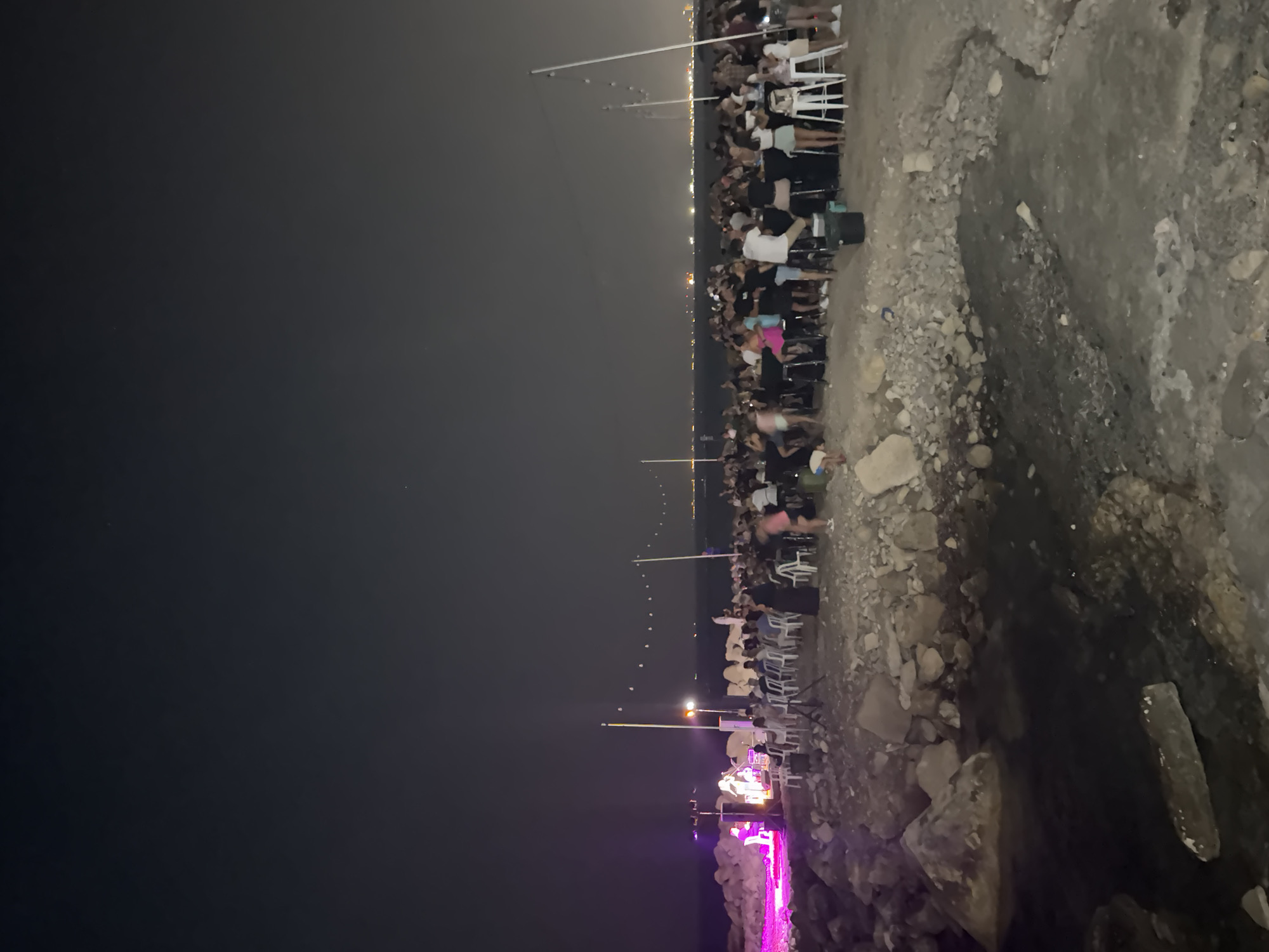At night, on a rocky shore in Agios Tychona Community, Cyprus, a crowd of people gathers for an outdoor event. The scene is illuminated by artificial lights, including purple stage lights on the left and string lights across the sky. In the foreground, the rocky terrain slopes down to the water's edge. A stage with performers is visible on the left, with an audience seated in chairs in front of it. Further to the right, a large group of people stand and mingle around tables set up on the rocks. The background shows a dark sea with a distant city skyline lit up along the horizon. The atmosphere appears to be festive and lively.