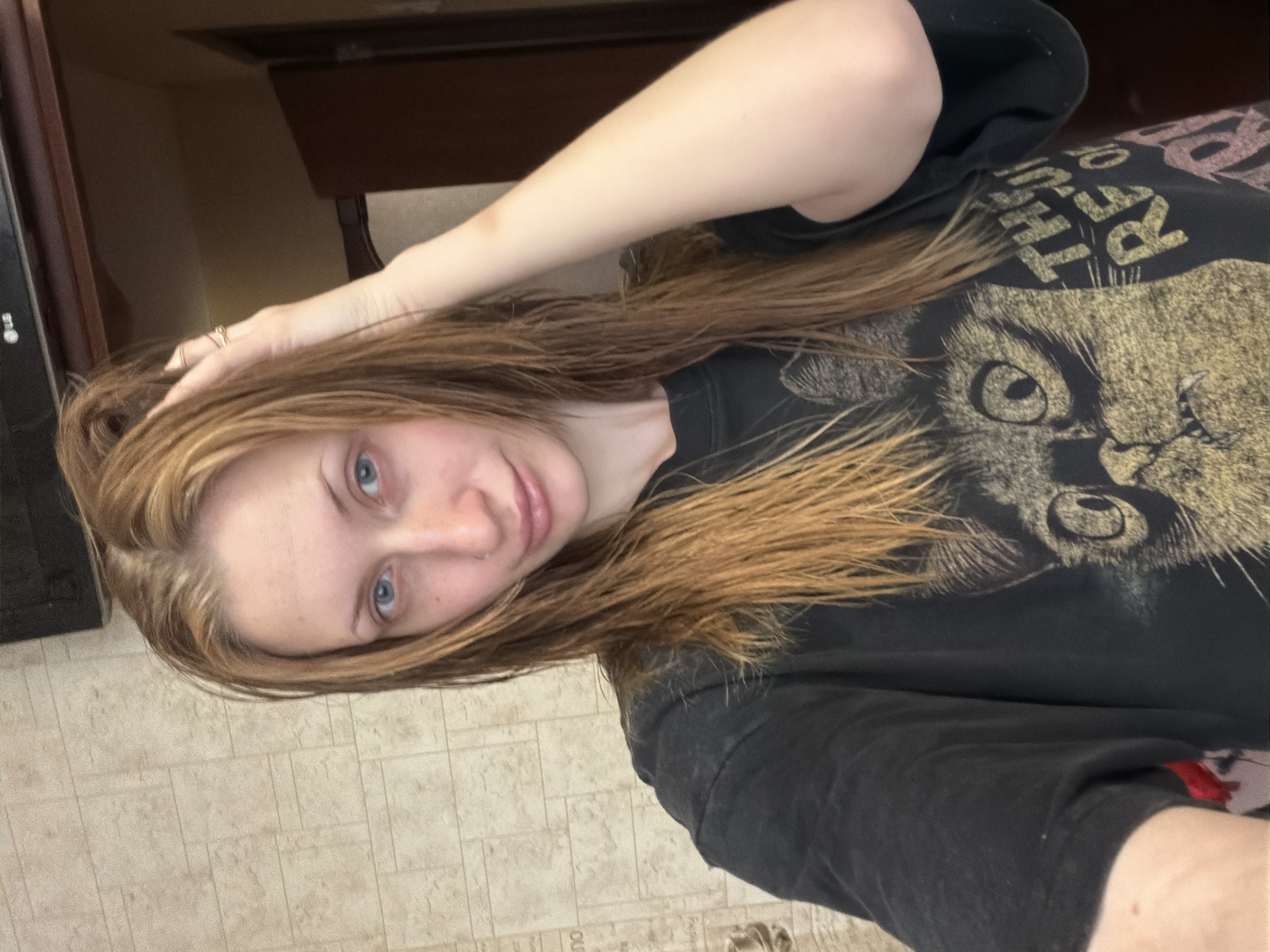This is a selfie taken by a young woman with light brown hair and blue eyes. She is wearing a black t-shirt with a graphic print of a cat's face and text that appears to say "THE CAT" in large, bold letters. The text is slightly obscured by her hair. The woman has her right hand raised, touching her hair and head. She is looking directly at the camera with a slight smile.
The setting appears to be indoors, possibly a home or a hotel room, given the presence of what looks like a dresser or wardrobe in the background and a tiled floor. The lighting suggests it is daytime. The woman's hair looks slightly damp, perhaps after a shower. There are no strong indicators of emotion other than a relaxed and pleasant demeanor. No weather cues are visible. The location context of Saint Petersburg, Russia, does not provide any specific visual cues within the image itself.