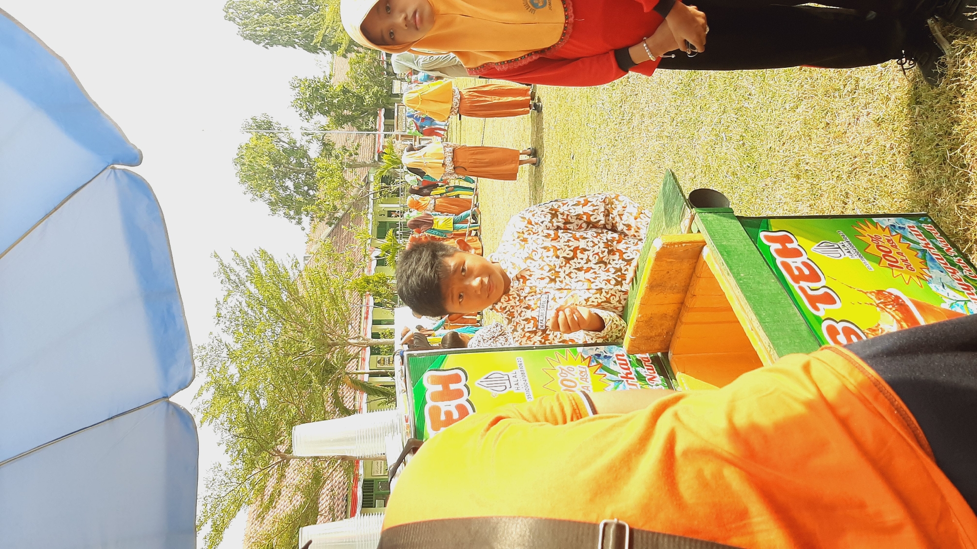 In Sumberjo, Indonesia, an outdoor scene unfolds, likely at a school or community event. A young boy, wearing a patterned shirt and a name tag reading "M. FAJAR AKBAR SAPUL", stands in front of a green and yellow beverage cart advertising "S TEH" (Ice Tea) and "100% GULA ASLI" (100% Original Sugar). He holds a small item in his right hand. To his right, a young girl in a red shirt and yellow hijab looks towards the viewer. Behind them, other children in similar attire are visible, some in orange and yellow outfits, suggesting a group or uniform. The background features buildings with red tile roofs and trees, under a large, light blue umbrella providing shade. The ground is covered in dry grass, indicative of a warm climate. The focus of the image is on the children and the beverage cart, suggesting a moment of interaction or a sales opportunity at the event.