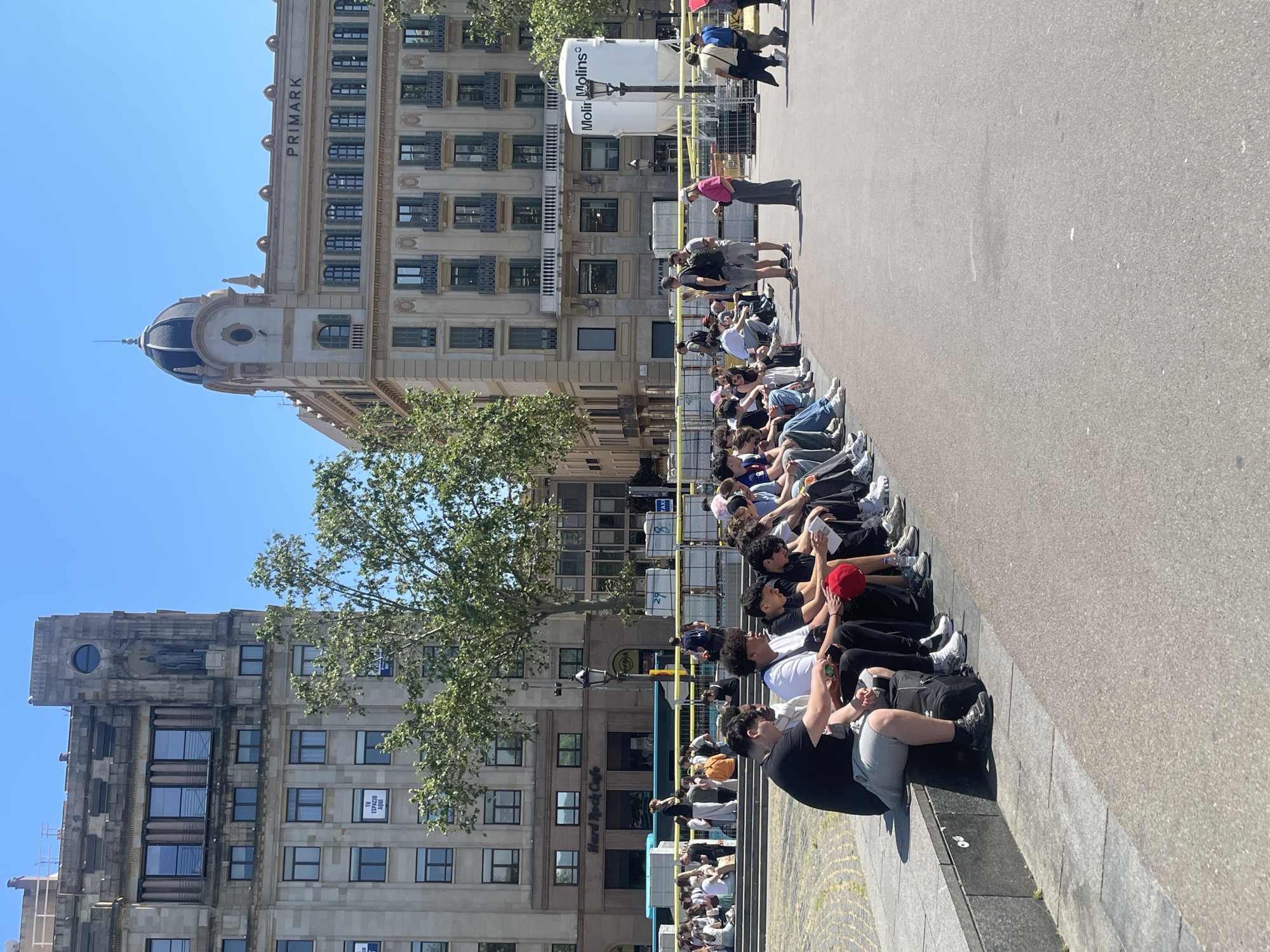 The image captures a vibrant urban street scene in Barcelona, Spain, under a clear, bright blue sky, indicating daytime, likely morning or early afternoon. The central focus is a long line of people, primarily young adults, seated on the edge of a stone ledge or steps that run parallel to a wide pavement or road. Most are facing to the right of the frame, patiently waiting; some are looking at their mobile phones, while others simply observe their surroundings.
In the background, grand multi-story buildings define the urban landscape. A prominent light-colored building features the word "PRIMARK" near its roofline and is adorned with a distinctive blue-domed turret. Further down the street, another building displays the "Hard Rock Cafe" logo. A lush green tree stands between these structures.
Several white cylindrical street structures, possibly advertising or utility boxes, are visible along the street, one bearing the text "Molini Molins". A yellow safety tape or band runs along the edge of the pavement. Another building on the left side of the frame shows a sign in a window that reads "EN ESTADO 100%," and a partial "TAXI" sign can be seen further back. The scene suggests a busy public area, with people gathered in anticipation.