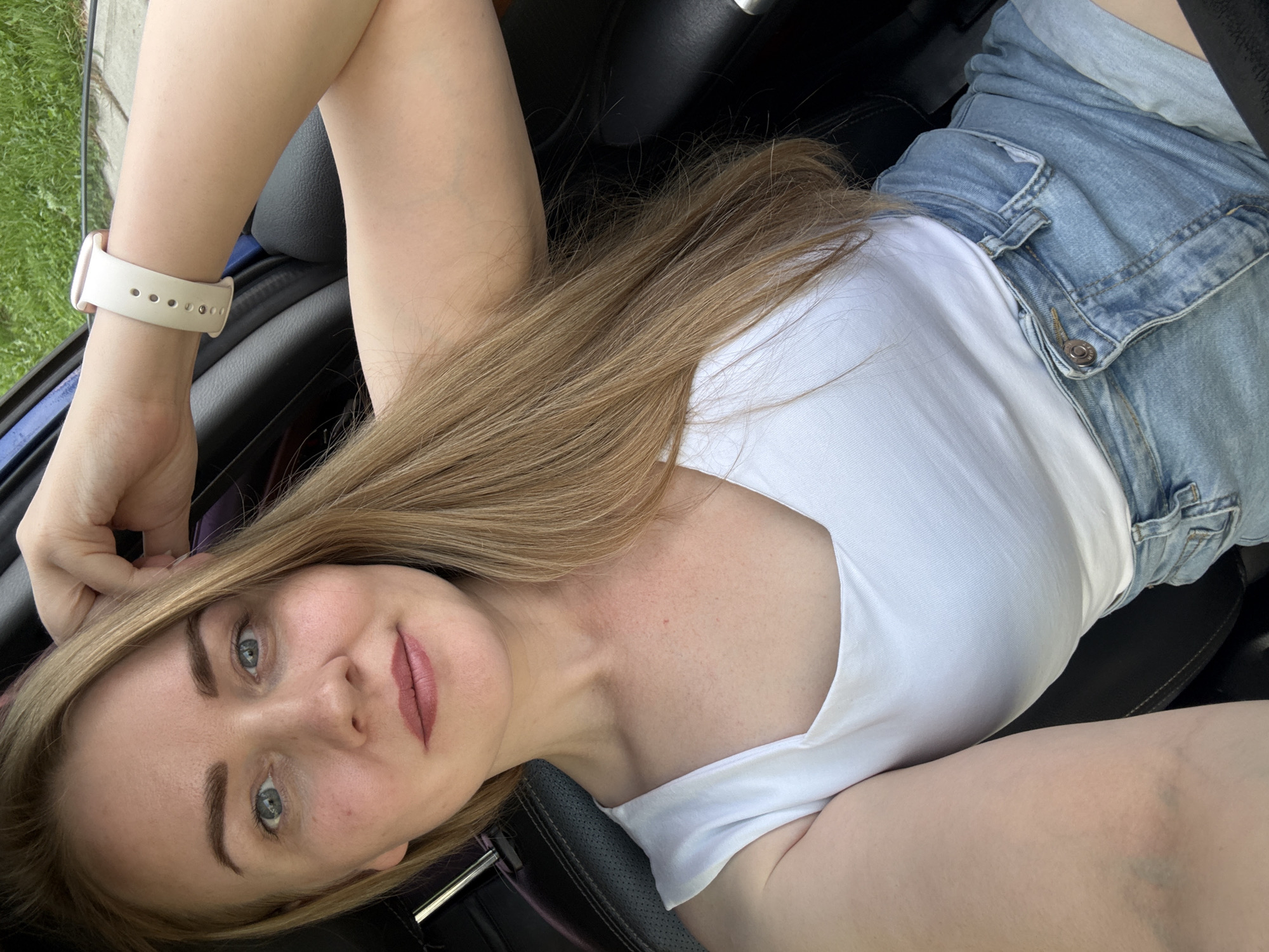 A woman with light blonde hair, wearing a white t-shirt and light blue denim shorts, is lying inside a vehicle. She is wearing a white smartwatch on her left wrist. The background shows a blurred view of green foliage, likely from a roadside. The location, Chaikovsky, Russia, is not discernible from the image alone.