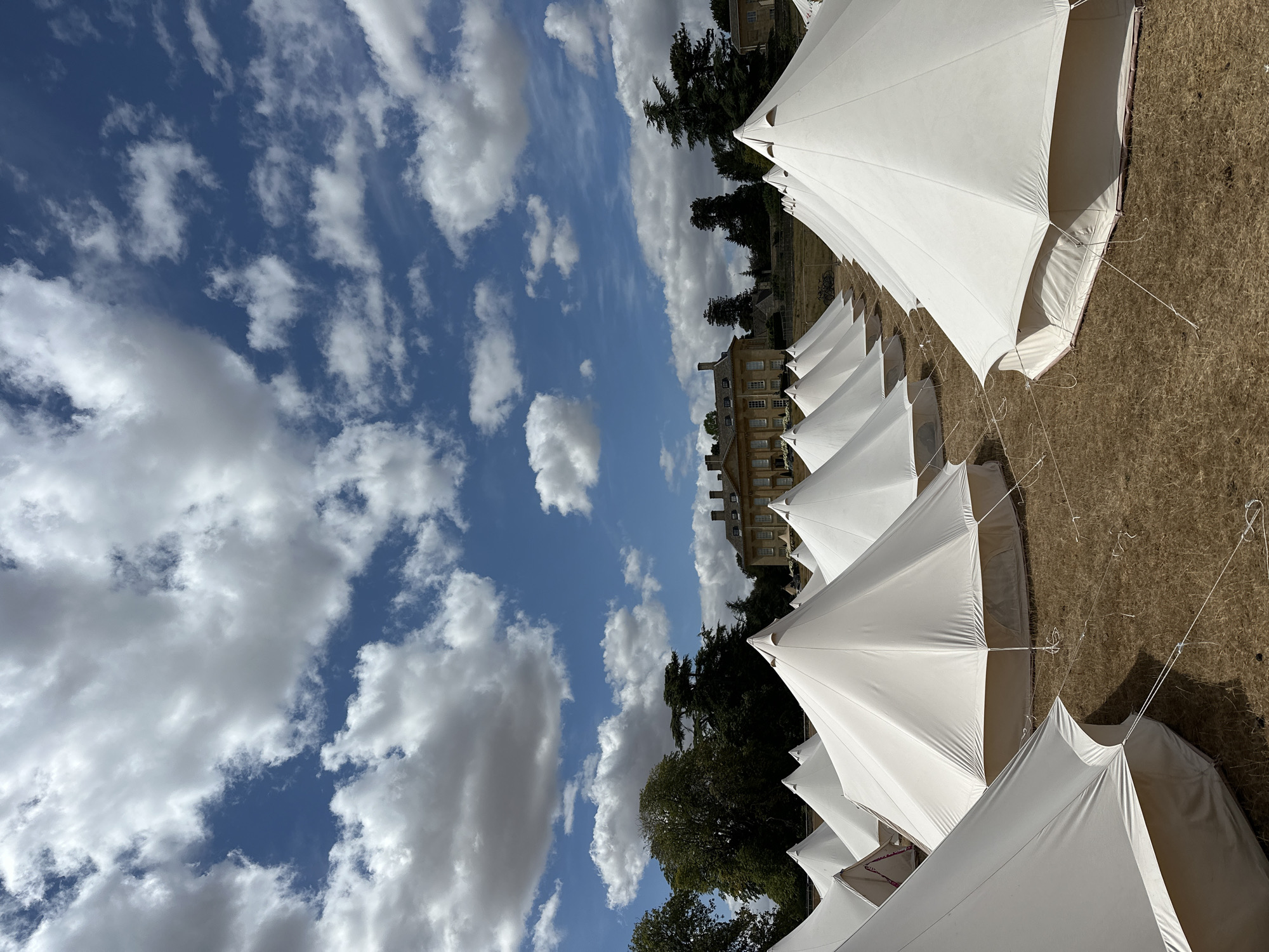 The image shows a large field dotted with numerous white bell tents, arranged in rows. The tents are conical in shape with fabric walls extending to the ground, secured by ropes and stakes. In the background, a grand, traditional building with a symmetrical facade, multiple windows, and a tiled roof stands amidst lush green trees. The sky above is a vibrant blue, populated by fluffy white clouds. The ground appears to be dry grass. The scene suggests an outdoor event or glamping site.