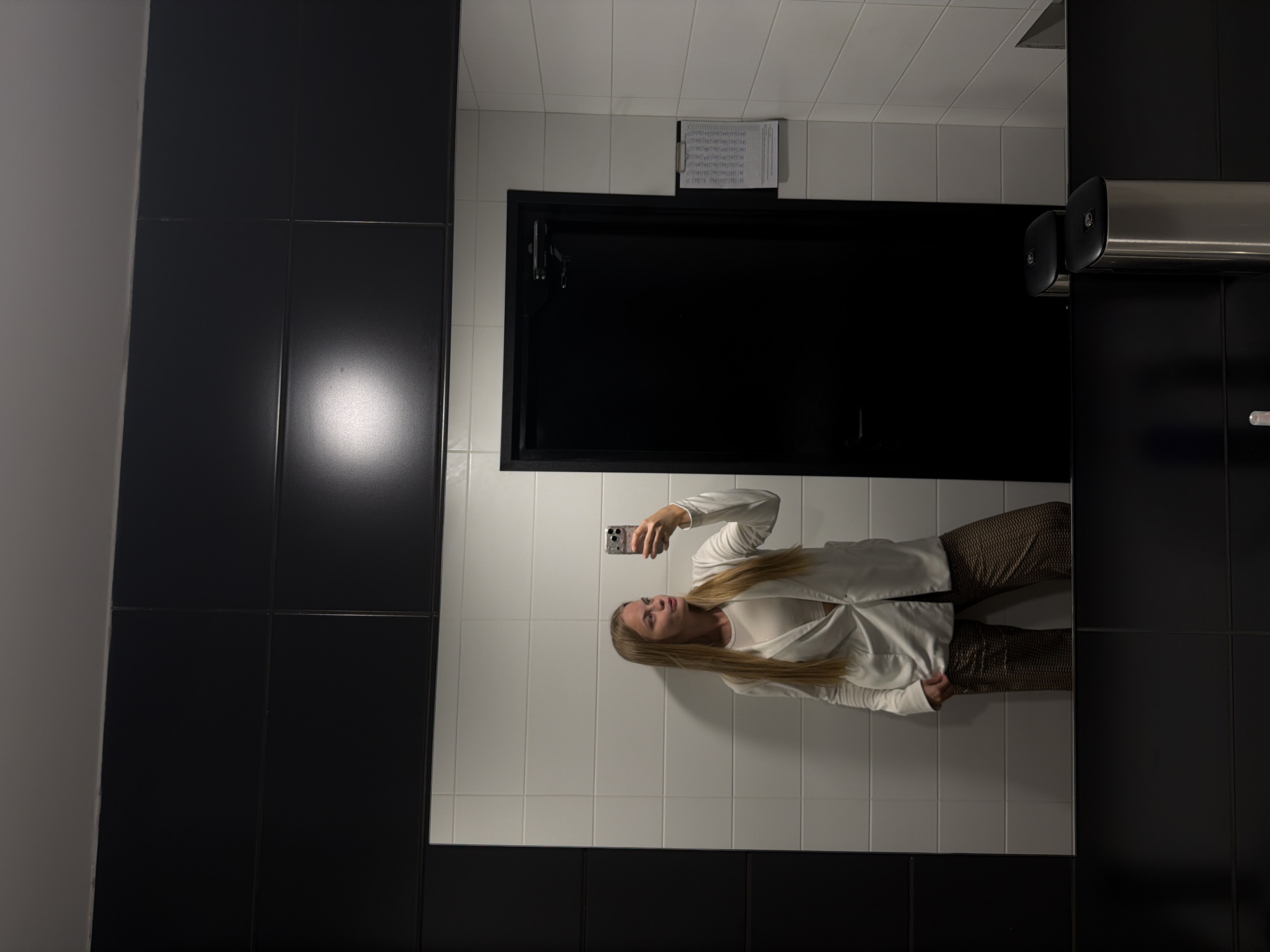 A woman is taking a mirror selfie in a bathroom. The bathroom has white tiled walls and black tiled sections on the left and right. There's a black cabinet above which a sheet of paper with text is mounted. A metal hand dryer is on the right wall. The woman is wearing a white shirt and patterned pants. The location is likely Izhevsk, Russia.