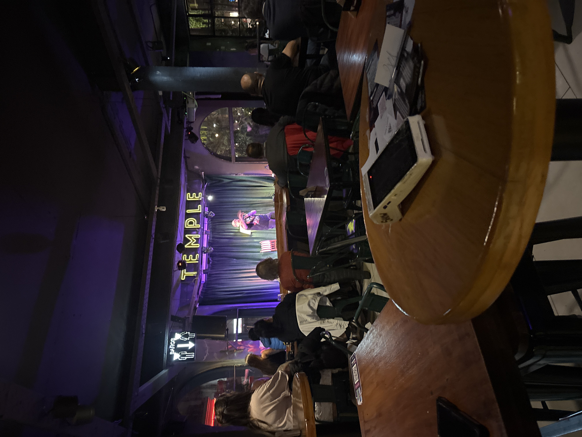 This medium shot captures an indoor evening scene within a dimly lit music venue or bar. The perspective is from a slightly elevated position, looking towards a stage where a live performance is underway.
**Subjects:**
Numerous people are seated at wooden tables, facing the stage. Most are seen from behind, dressed in casual attire. In the foreground, a light-colored wooden table occupies the right side of the frame, with a mobile device (possibly a portable game console or a power bank) and some papers visible on its surface. Further back, audience members are seated at smaller, darker tables and chairs. On the stage, a single performer, likely a musician or singer, is holding a microphone and addressing the audience.
**Activity or event taking place:**
A live musical performance or a stand-up act is taking place on the stage. The audience is gathered to watch and listen, indicating a social event at a entertainment venue.
**Notable details:**
The venue is characterized by its dark ambiance, illuminated primarily by stage lights and neon signs. A strong purple hue emanates from the stage area, lighting up the walls and ceiling. Behind the performer, large purple drapes or curtains serve as a backdrop. Above the stage, neon yellow letters clearly spell out "TEMPLE". To the left of the stage, another neon sign with white light indicates "BAÑOS" (restrooms) with an arrow pointing upwards. The tables and chairs suggest a casual setting for enjoying drinks and entertainment. The time of day is definitively evening or night due to the artificial lighting. The overall atmosphere appears intimate and relaxed.
**Visible text:**
- "TEMPLE" (in yellow neon letters above the stage)
- "BAÑOS" (in white neon letters, pointing upwards, for restrooms)